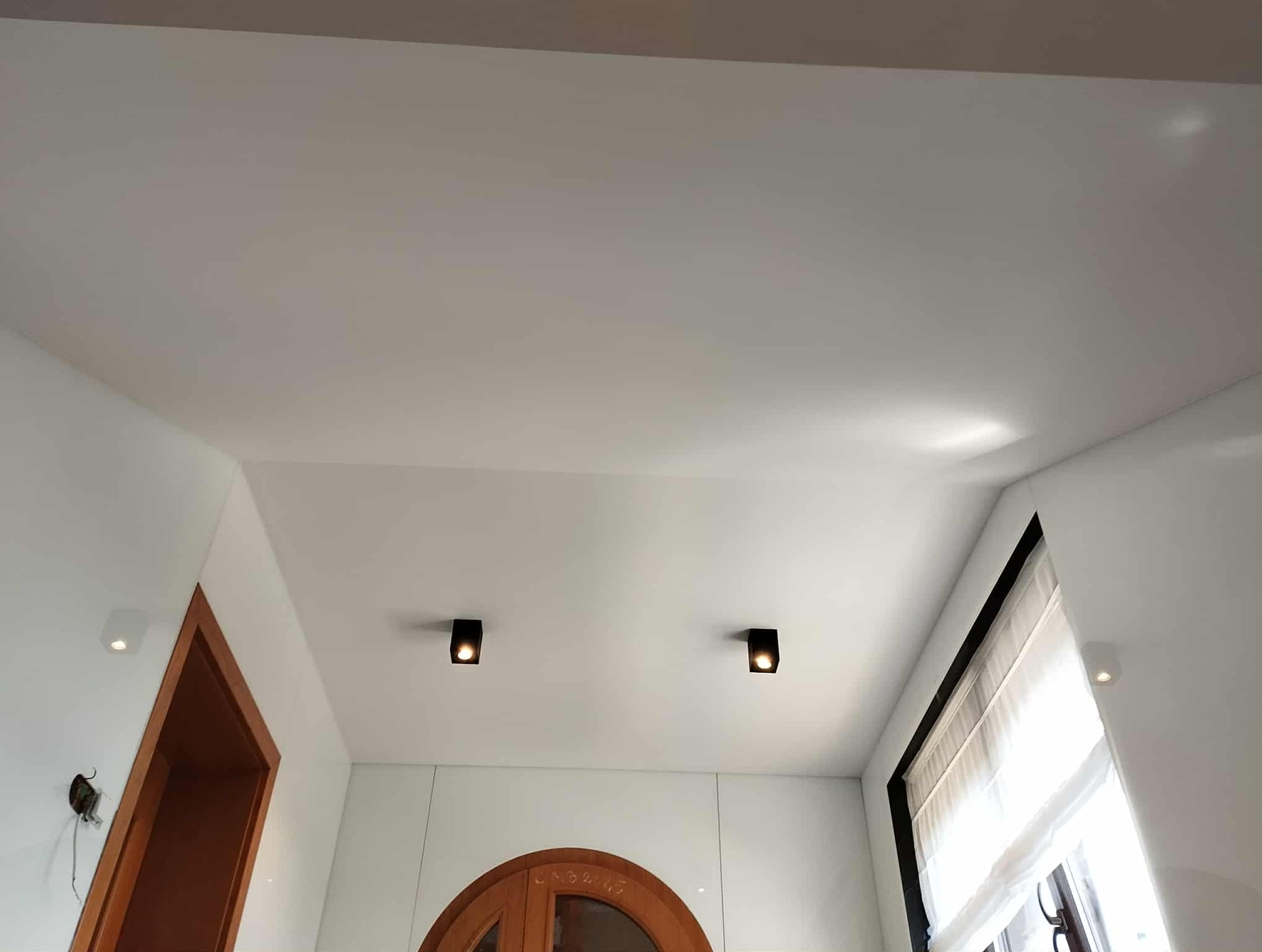 Biały sufit napinany z dwoma czarnymi, punktowymi lampami LED. Widoczne fragmenty okna z roletą rzymską i drewnianych drzwi z łukowym nadprożem.
