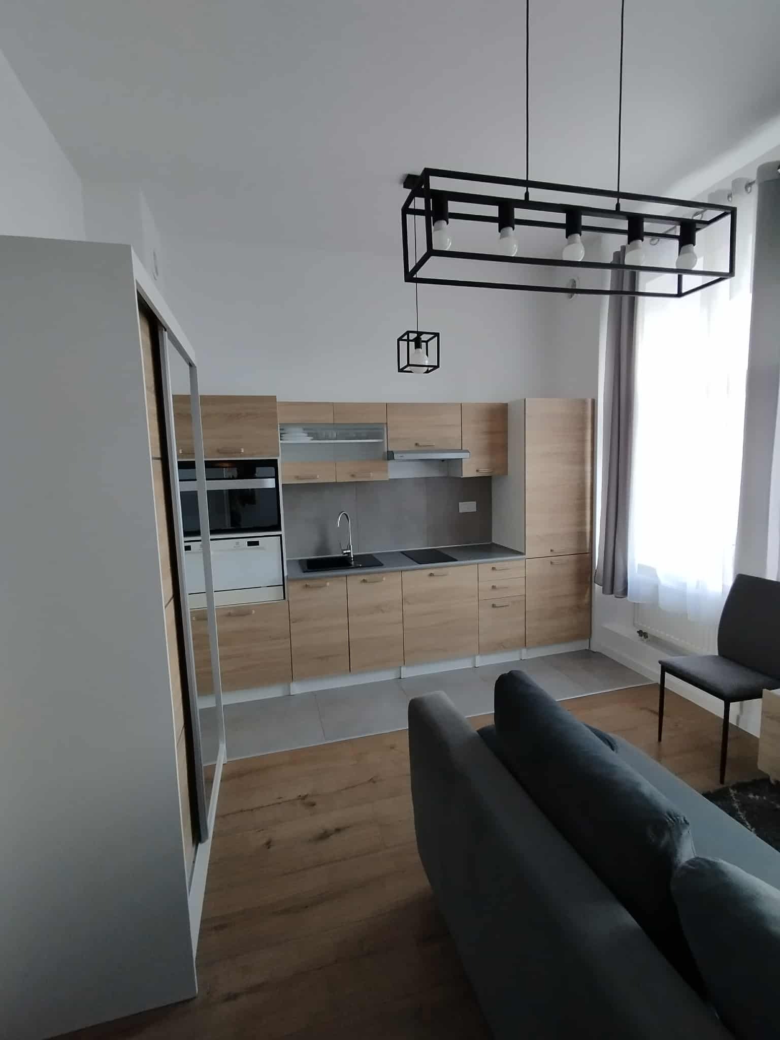 Jasna kuchnia z drewnianymi frontami, szarym blatem i nowoczesnym oświetleniem. Widoczny fragment sofy i szafy wnękowej. Minimalistyczny design.