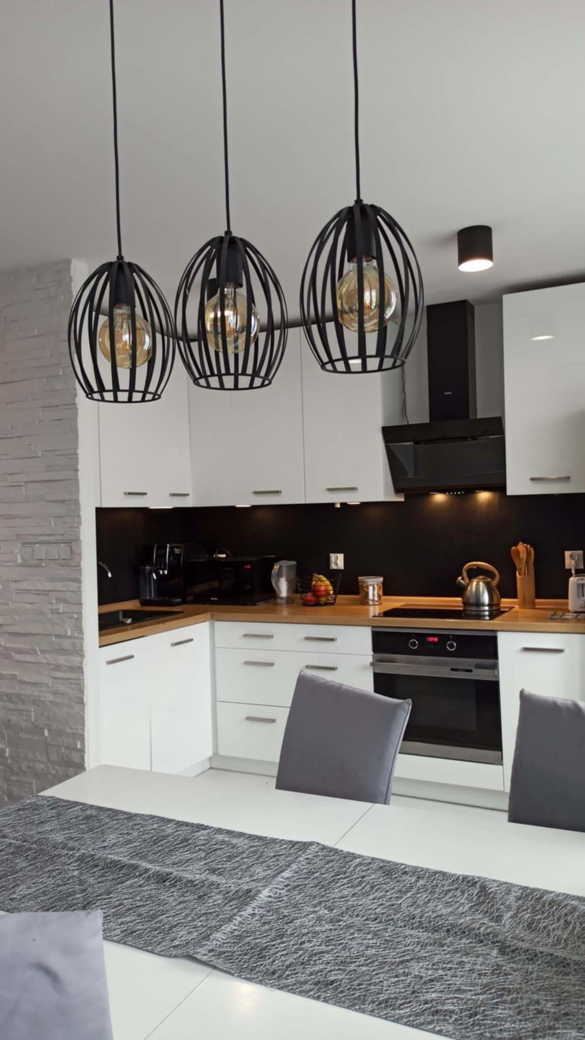 Nowoczesna kuchnia z białymi szafkami, czarnym panelem i trzema wiszącymi lampami w stylu industrialnym. Na blacie ekspres do kawy i czajnik.