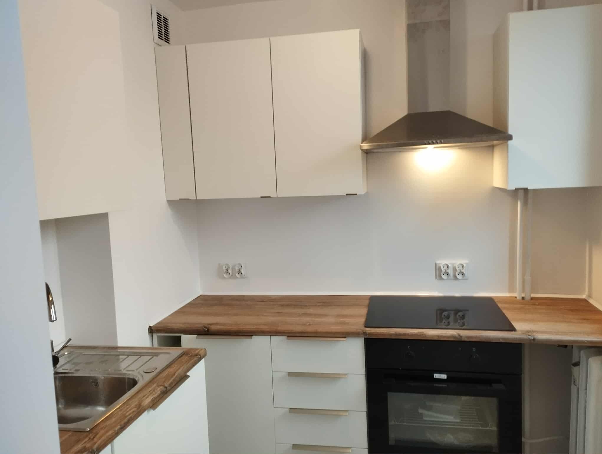 Nowoczesna kuchnia z białymi szafkami i drewnianym blatem. Czarna płyta indukcyjna i piekarnik w zabudowie. Minimalistyczny design z oświetleniem punktowym.