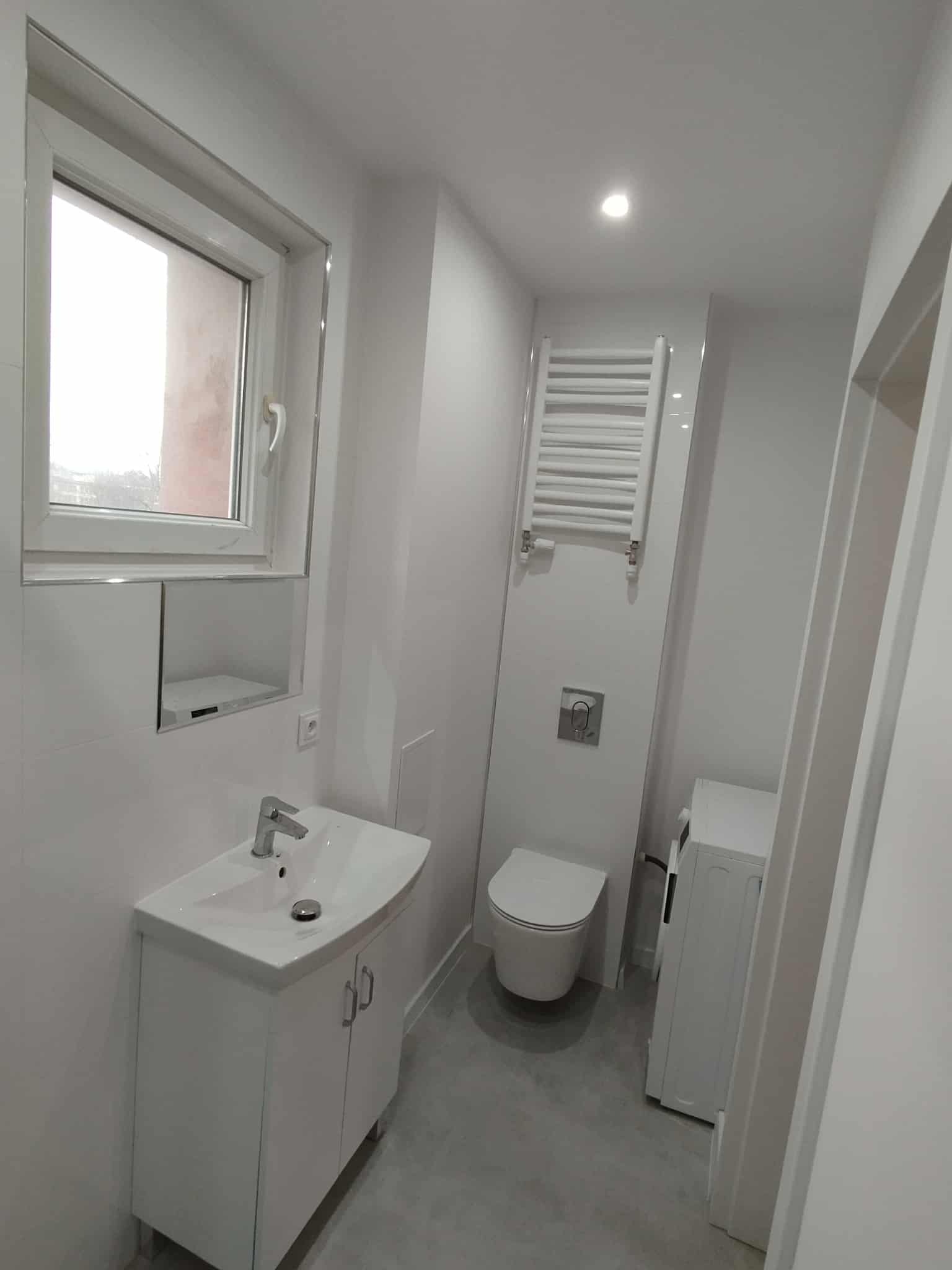 Jasna łazienka po remoncie: białe płytki, umywalka z szafką, toaleta, grzejnik drabinkowy i pralka. Minimalistyczny design z oknem i lustrem.