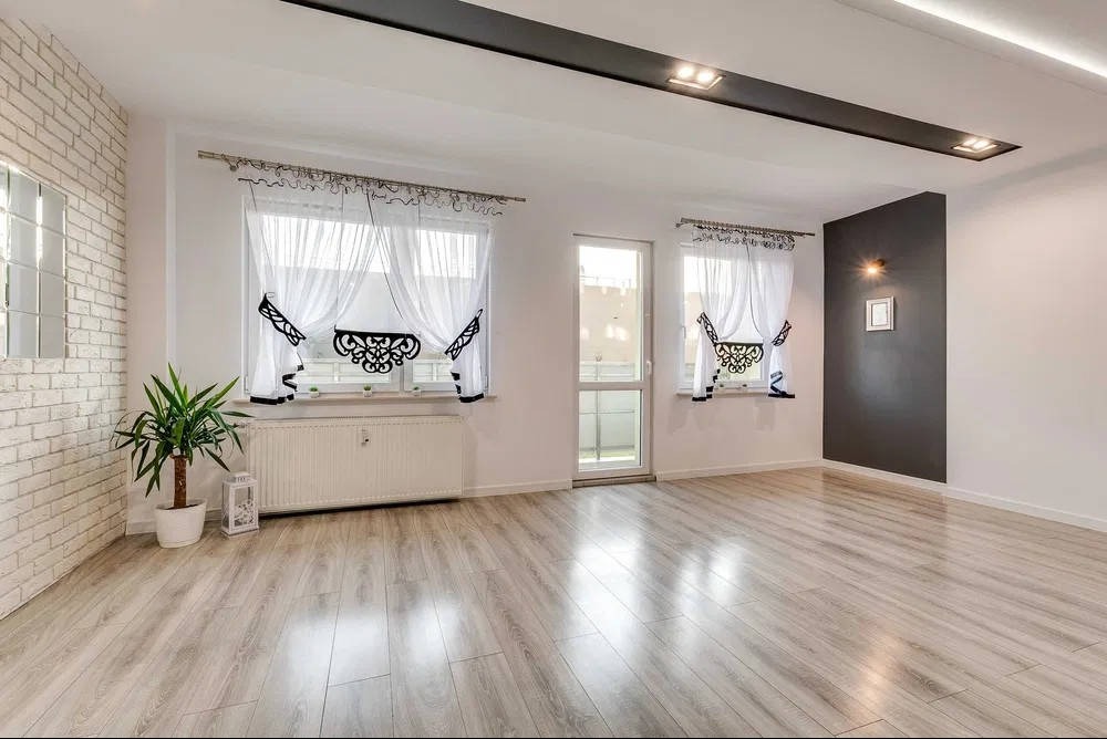 Jasne, przestronne wnętrze po remoncie: panele podłogowe, ceglana ściana, okna z dekoracyjnymi firanami, minimalistyczne oświetlenie sufitowe i ciemna ściana akcentująca.