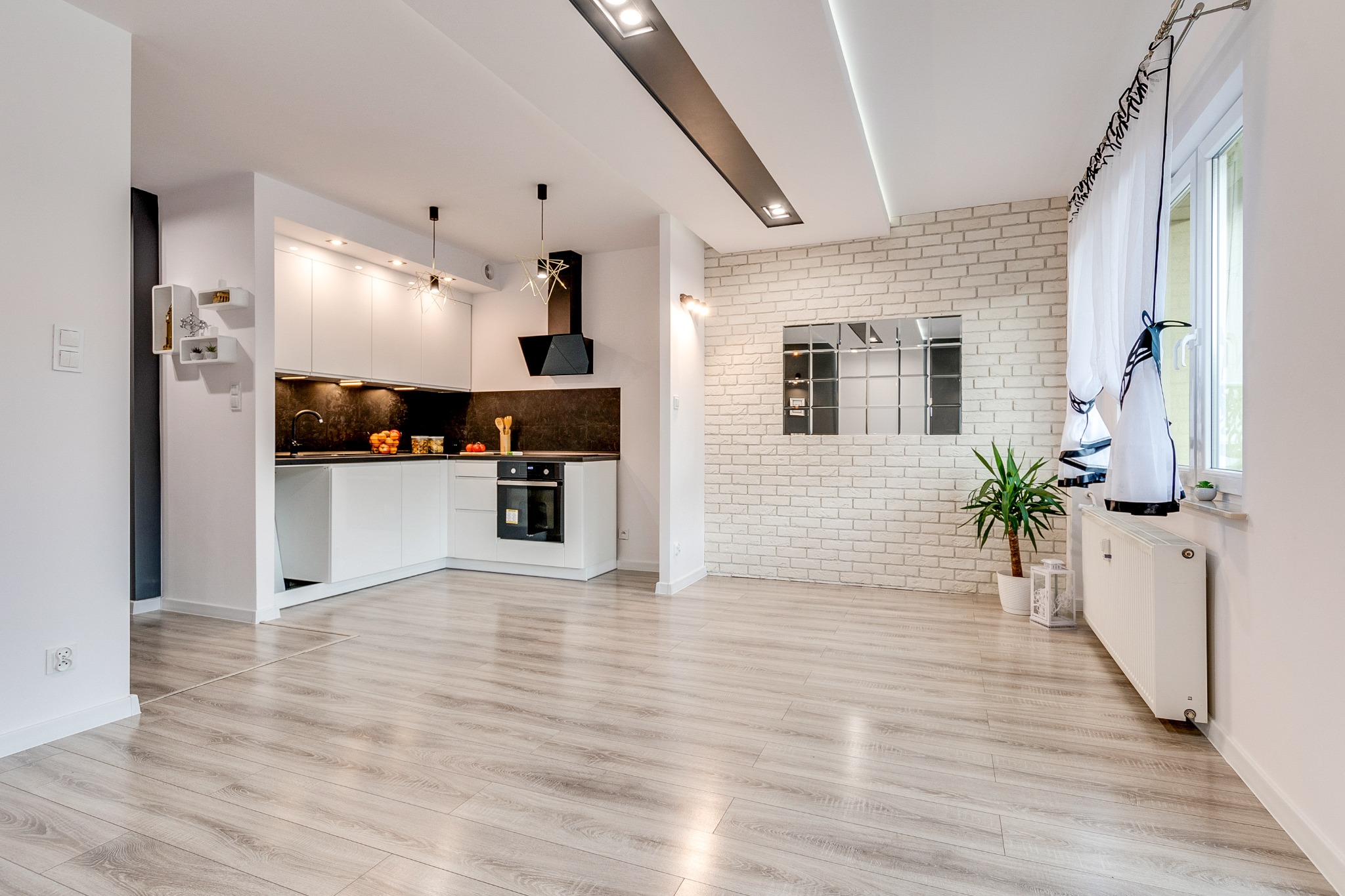 Jasne, przestronne wnętrze po remoncie: biała kuchnia z czarnym akcentem, ściana z białej cegły, drewniana podłoga i okno z firanką. Nowoczesny design.