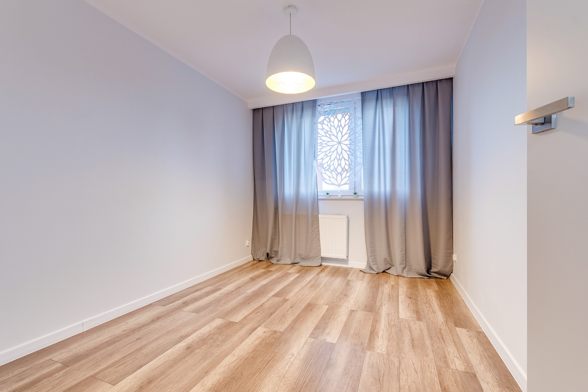 Jasne panele podłogowe w pokoju z białymi ścianami, szarymi zasłonami i ozdobnym ekranem okiennym. Nowoczesna lampa sufitowa i kinkiet dopełniają wystrój.