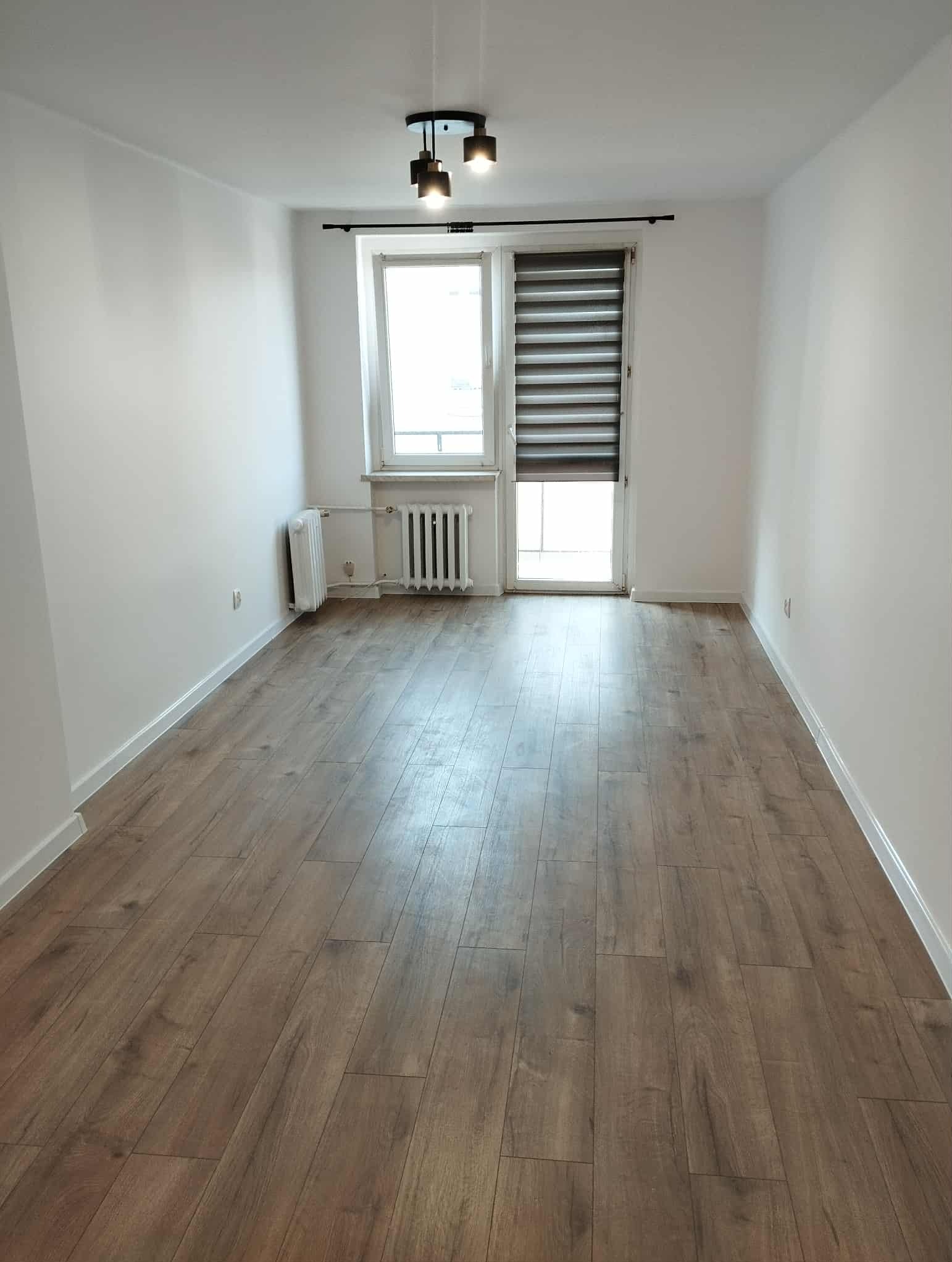 Jasne wnętrze po remoncie: drewnopodobna podłoga, białe ściany, okno z roletą dzień-noc i nowoczesna lampa sufitowa. Minimalistyczny design.