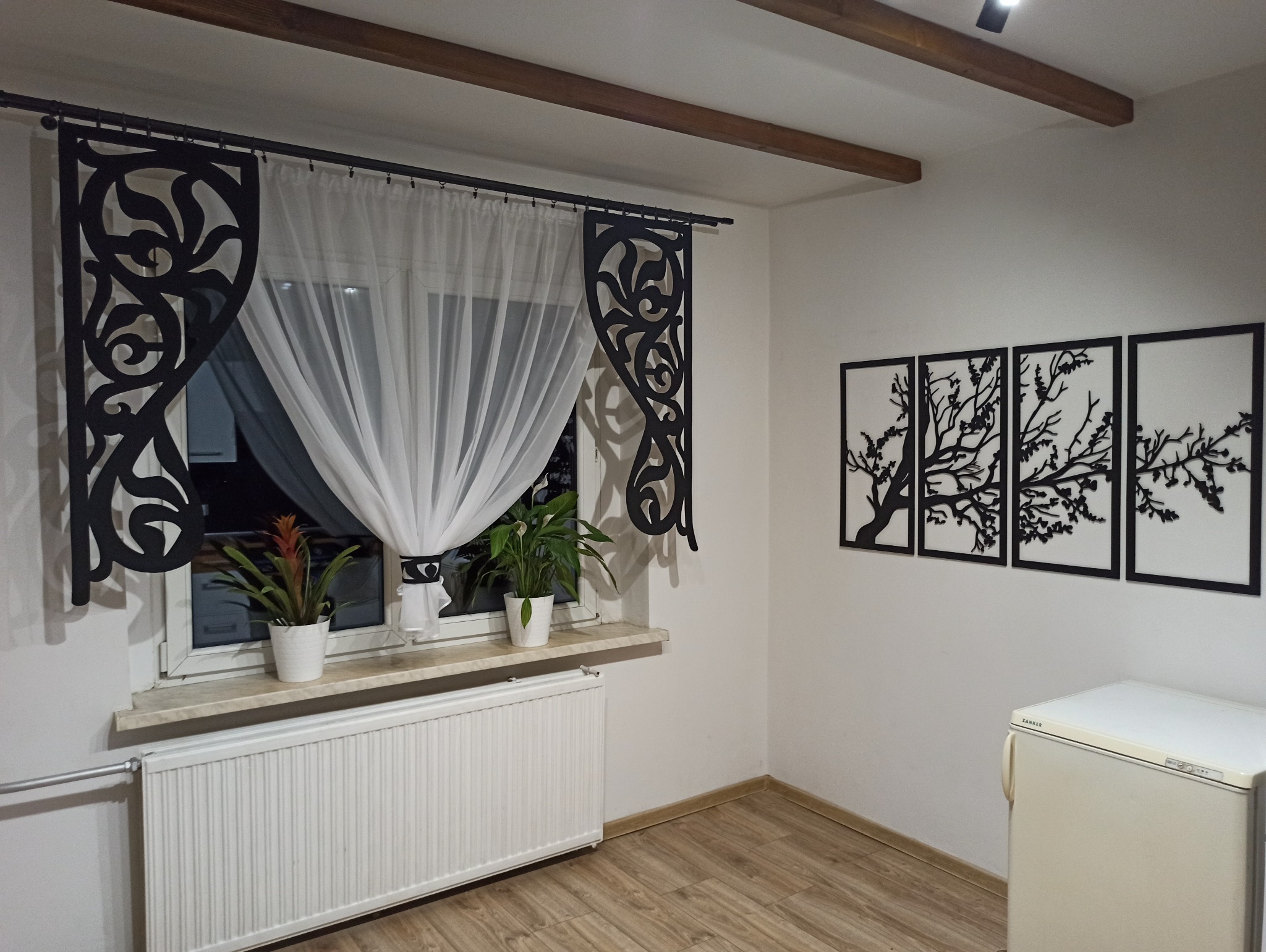 Nowoczesny wystrój wnętrza: okno z dekoracyjnymi panelami, firanka, rośliny w doniczkach na parapecie i cztery obrazy z motywem drzewa na ścianie.