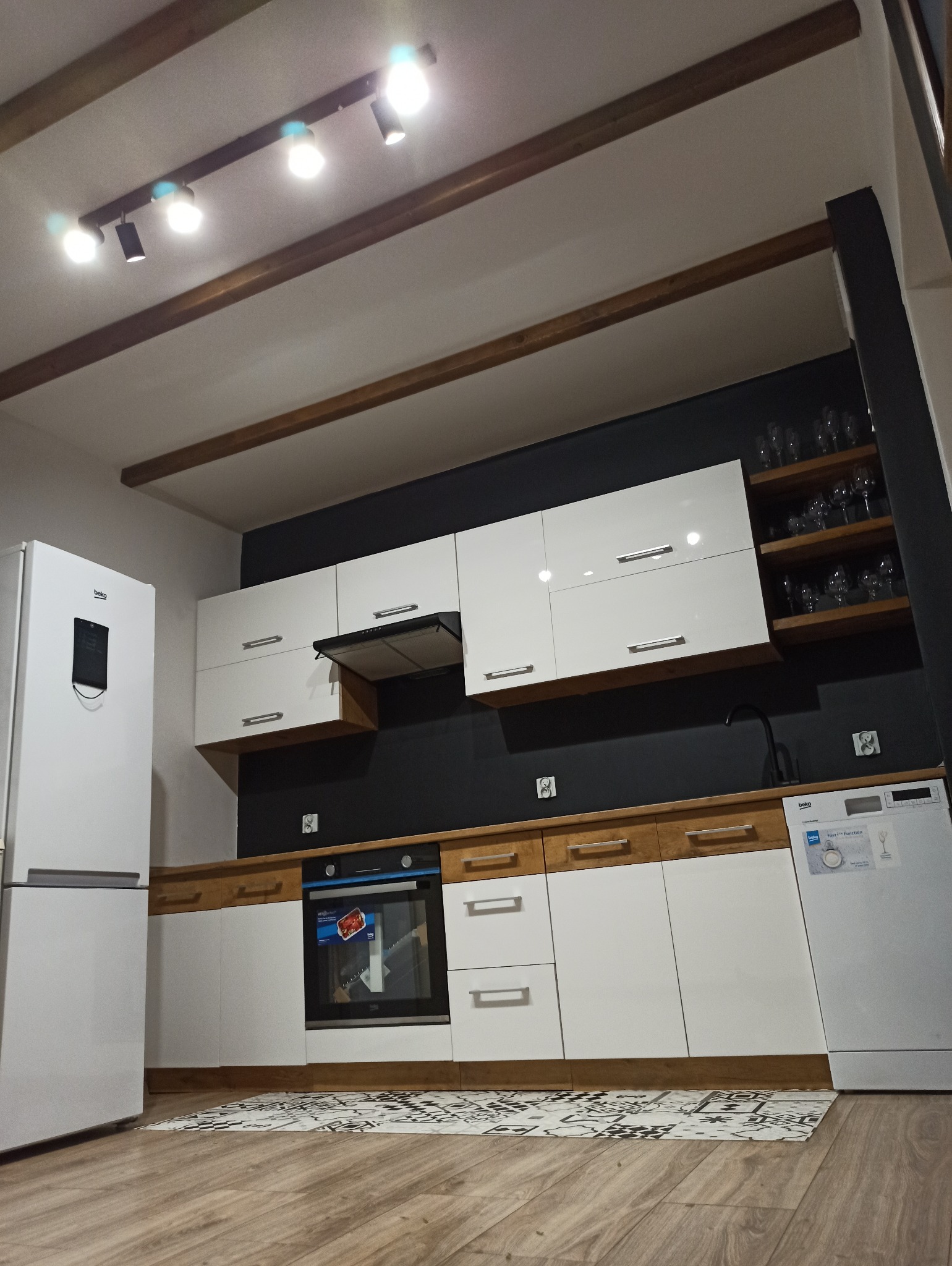 Nowoczesna kuchnia z białymi szafkami, drewnianymi blatami i czarną ścianą. Widoczne oświetlenie sufitowe i sprzęt AGD: lodówka, piekarnik, zmywarka. Podłoga drewniana.