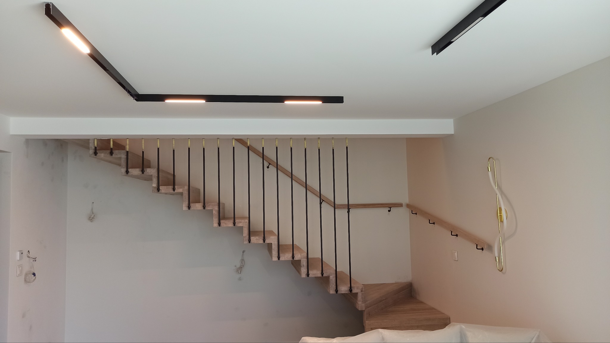 Nowoczesne schody z drewnianymi stopniami i czarnymi balustradami, oświetlone liniowymi lampami sufitowymi. Minimalistyczny design, jasne ściany.