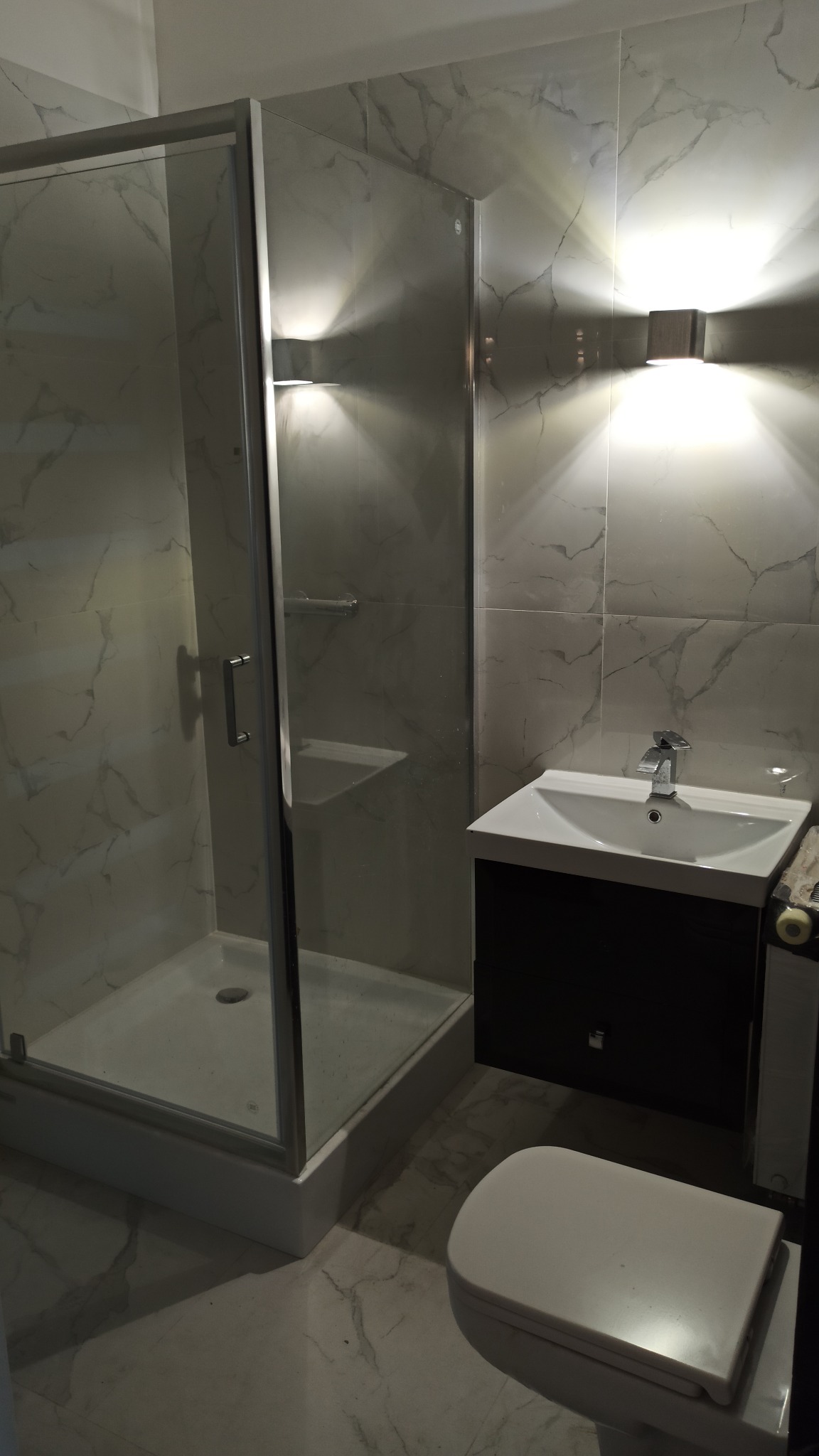 Nowoczesna łazienka z kabiną prysznicową, umywalką z szafką i toaletą. Marmurowe płytki ścienne i podłogowe. Oświetlenie punktowe nad umywalką.