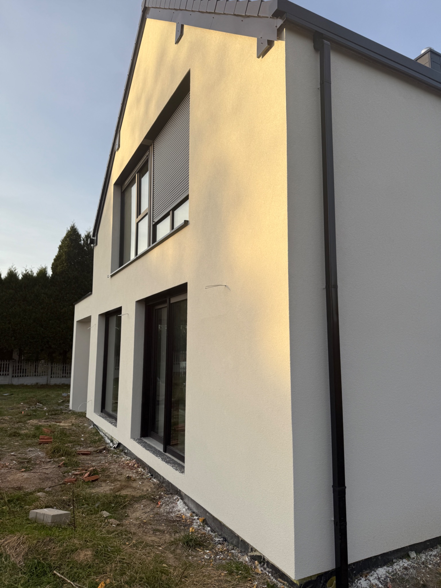 Boczny widok domu po ociepleniu elewacji, z widocznymi oknami z roletami, rynną i fragmentem terenu z resztkami materiałów budowlanych. Jasna elewacja w promieniach słońca.