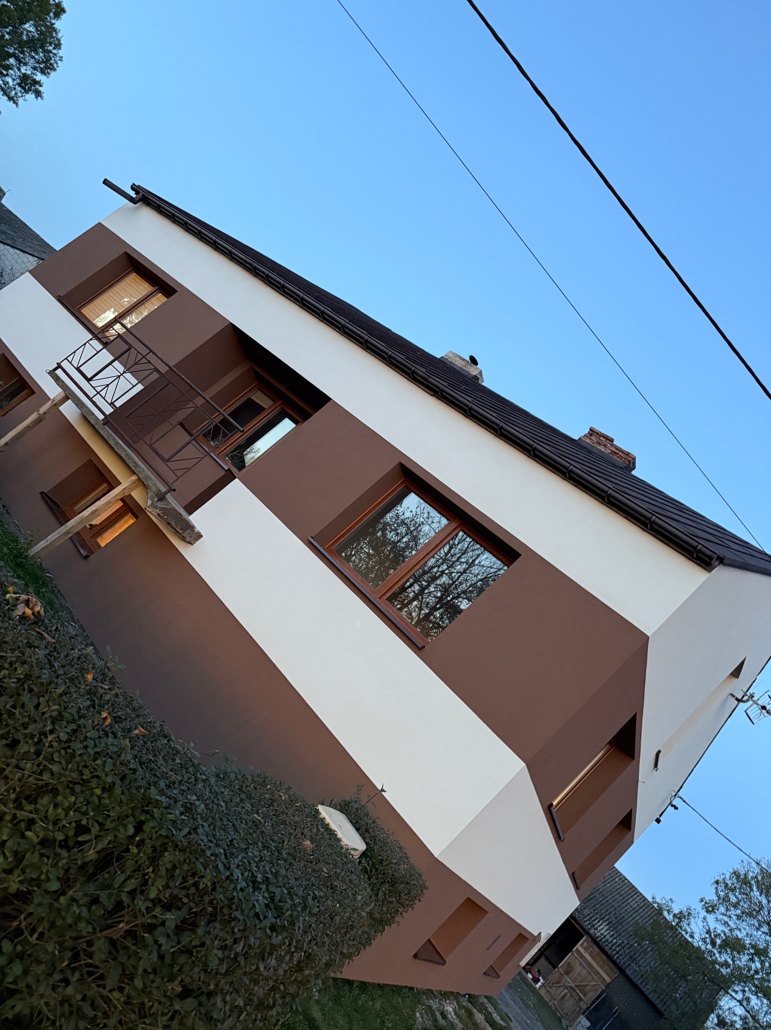 Dwukondygnacyjny dom z elewacją w odcieniach brązu i bieli, z balkonem i widocznymi oknami, na tle błękitnego nieba i zieleni. Ujęcie z dołu, podkreślające geometrię budynku.
