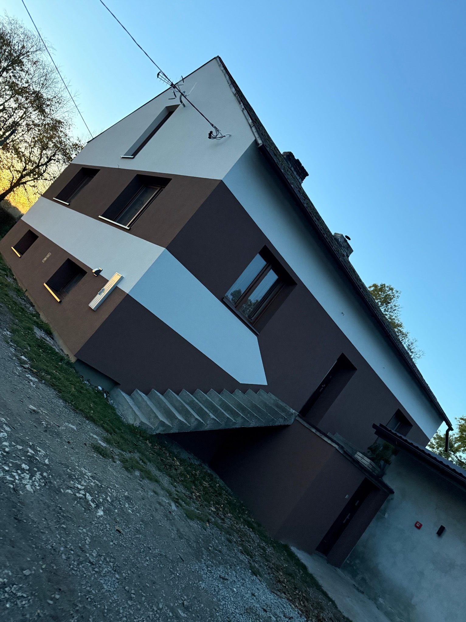 Dwukondygnacyjny dom z elewacją w geometryczne pasy brązu i bieli, widziany z dołu pod kątem, z widocznymi schodami wejściowymi i fragmentem podwórza.