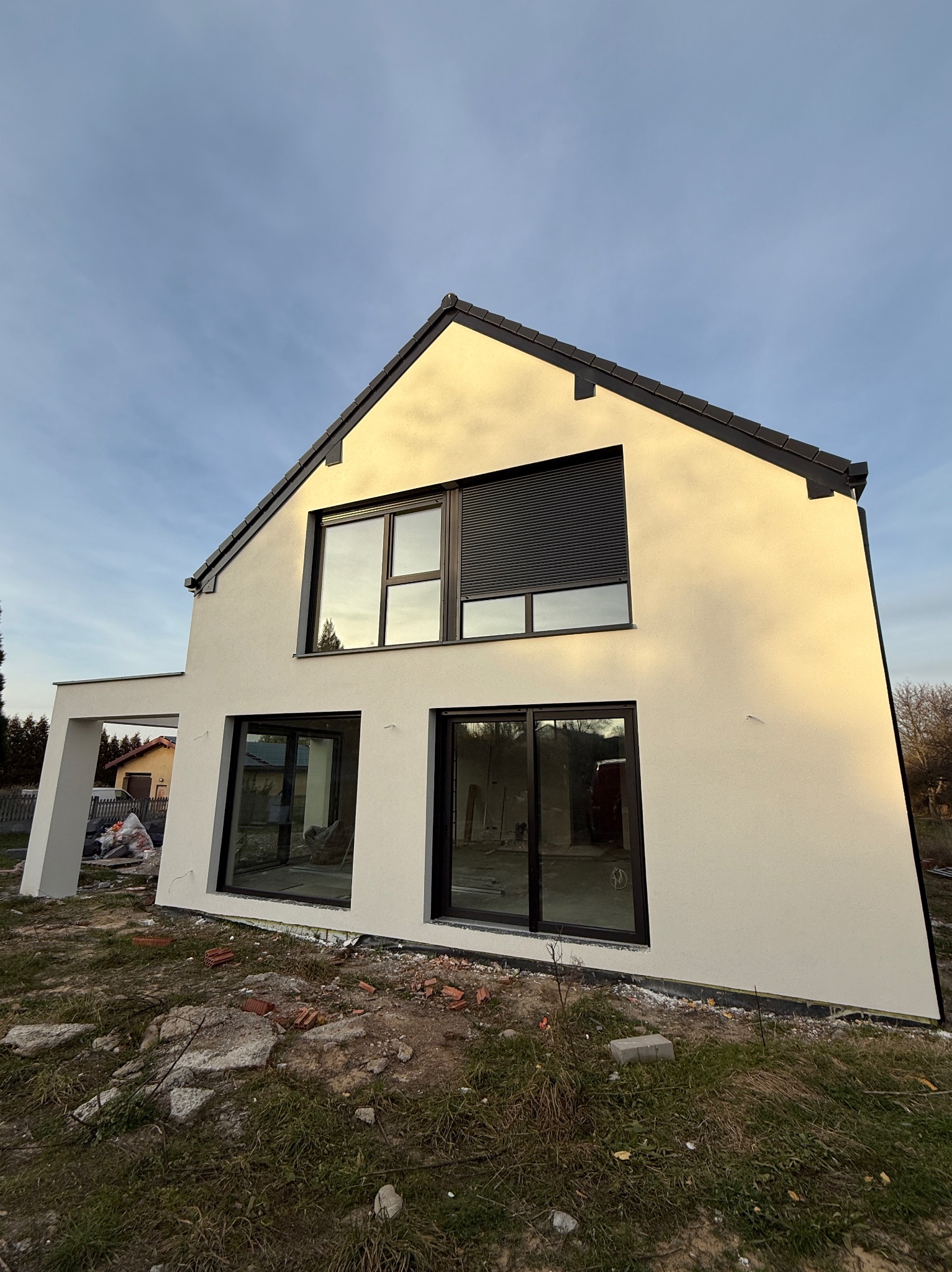 Nowoczesny dom jednorodzinny z jasną elewacją i ciemną stolarką okienną, widziany z perspektywy ogrodu. Widoczne fragmenty terenu po pracach budowlanych.