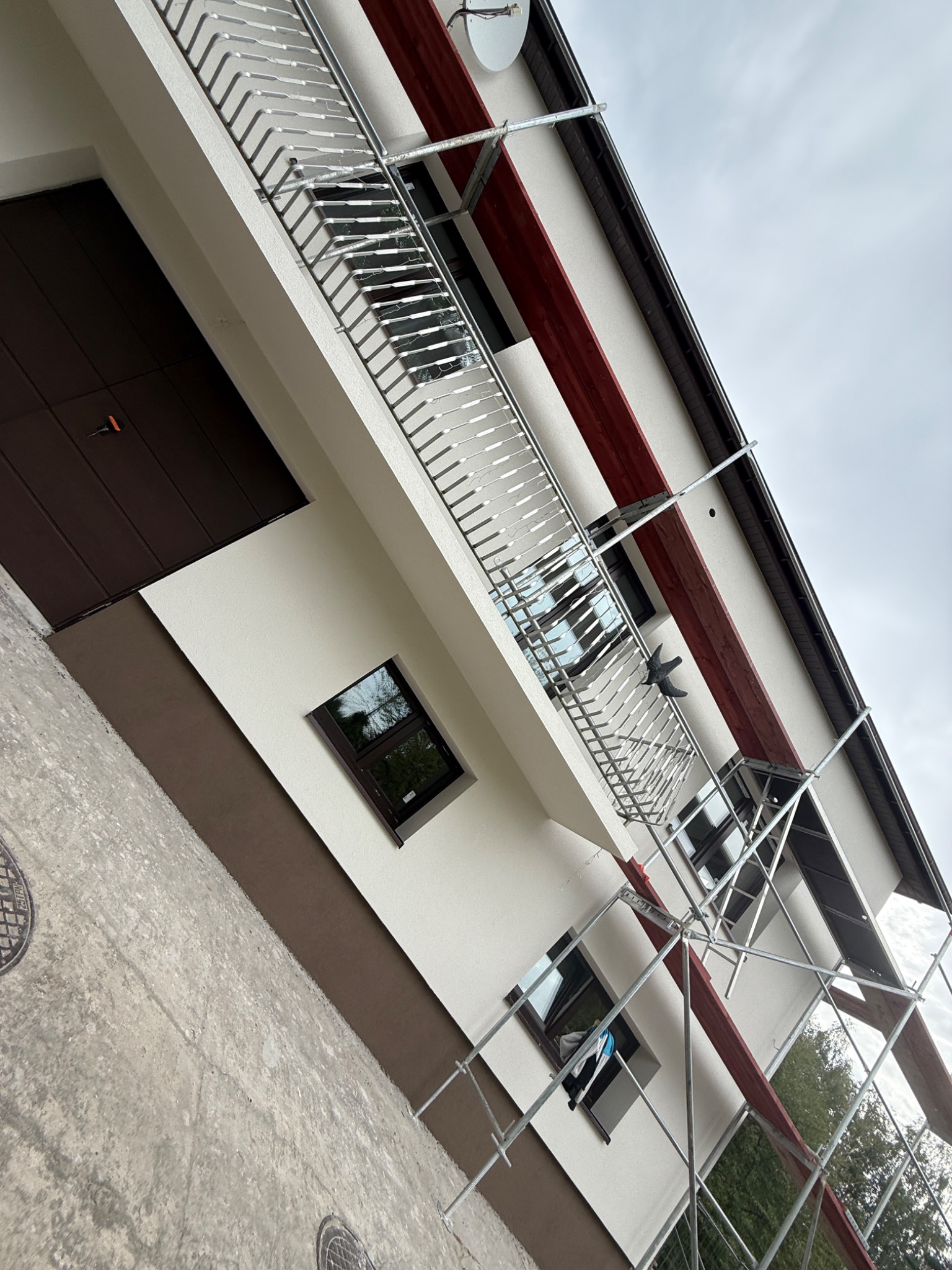 Świeżo pomalowana elewacja domu z balkonem, widoczna z dołu pod kątem. Na elewacji rusztowanie, brązowe akcenty i stalowa balustrada balkonu.