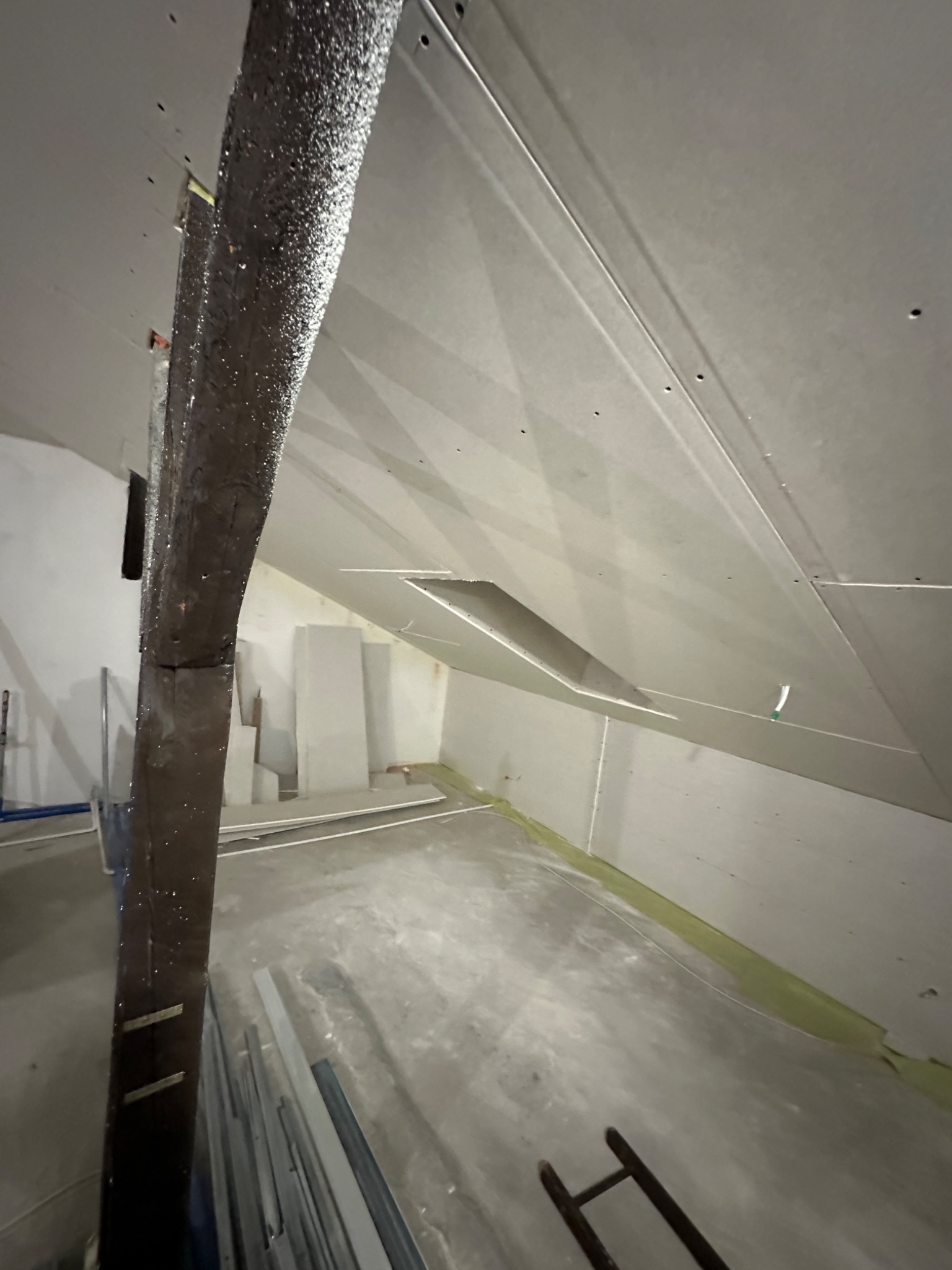 Poddasze w trakcie prac wykończeniowych: widoczne płyty gipsowo-kartonowe na skosach dachu, drewniany słup konstrukcyjny i materiały budowlane na podłodze.