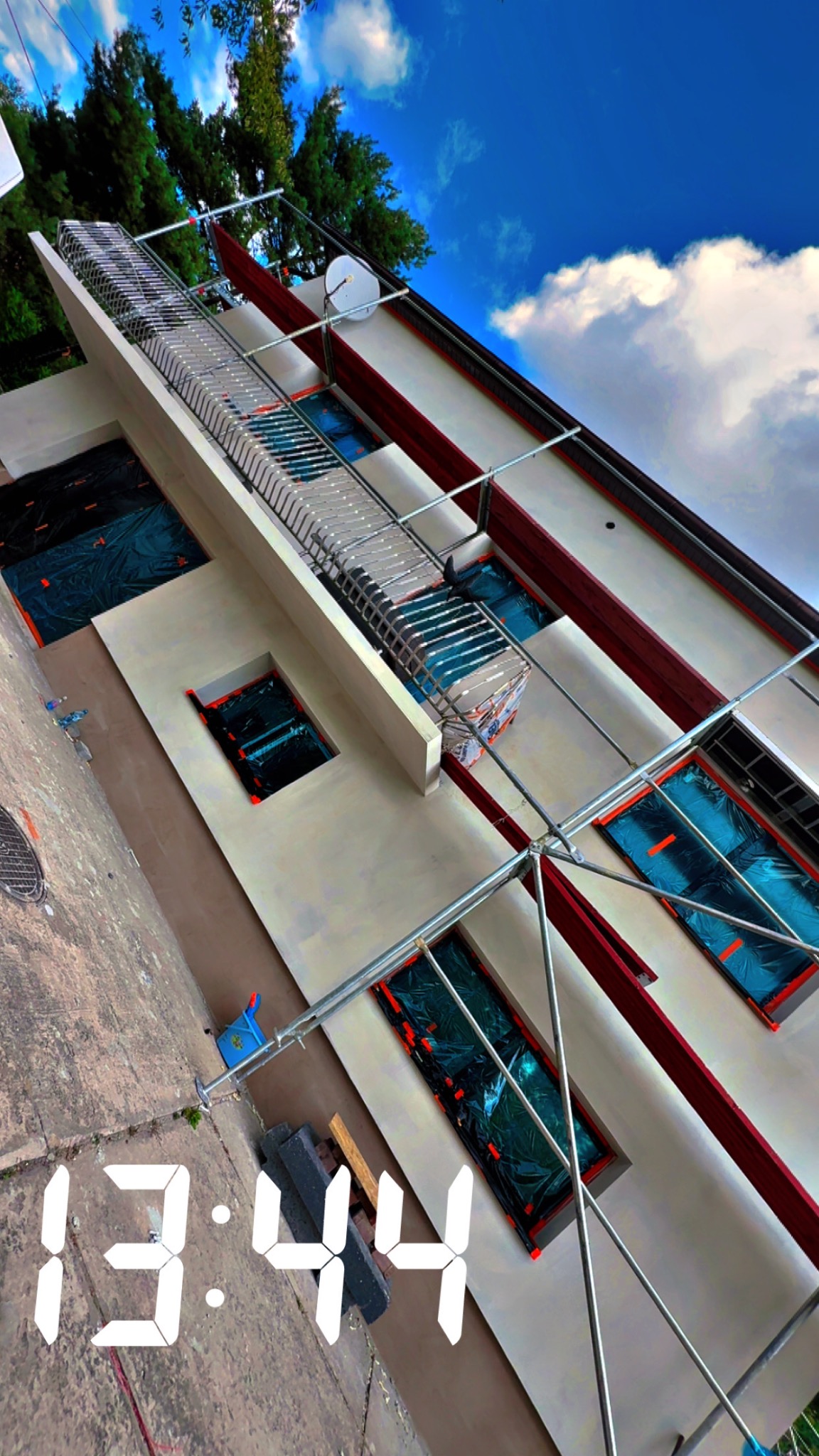 Elewacja budynku w trakcie prac: okna zabezpieczone folią, widoczne rusztowania. Świeżo otynkowana ściana w jasnym kolorze. Ujęcie z dołu, widoczne niebo.