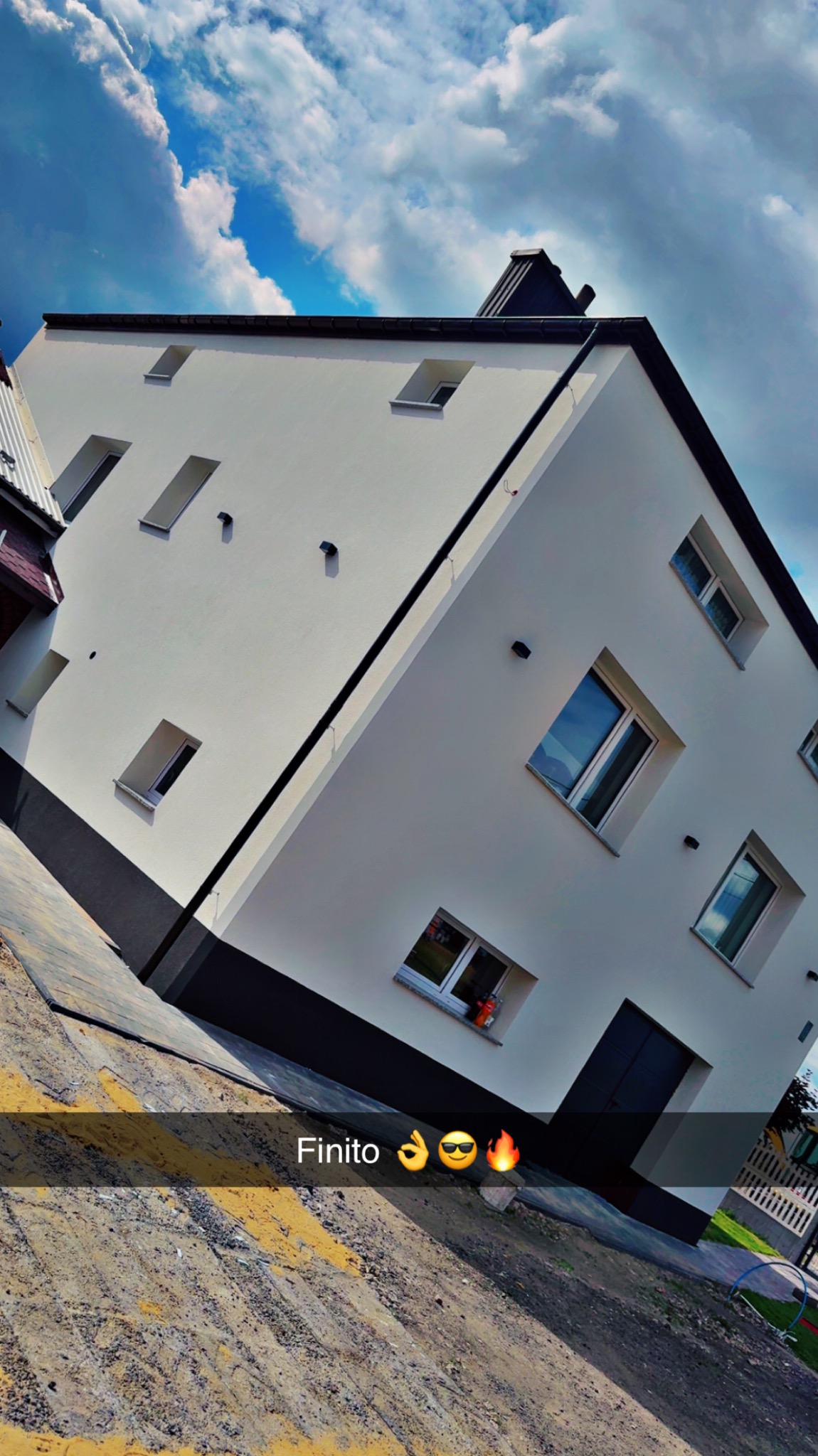 Biała elewacja budynku z czarnymi akcentami, widoczna z dołu pod kątem. Okna odbijają niebo. Na pierwszym planie fragment chodnika z żółtymi pasami.