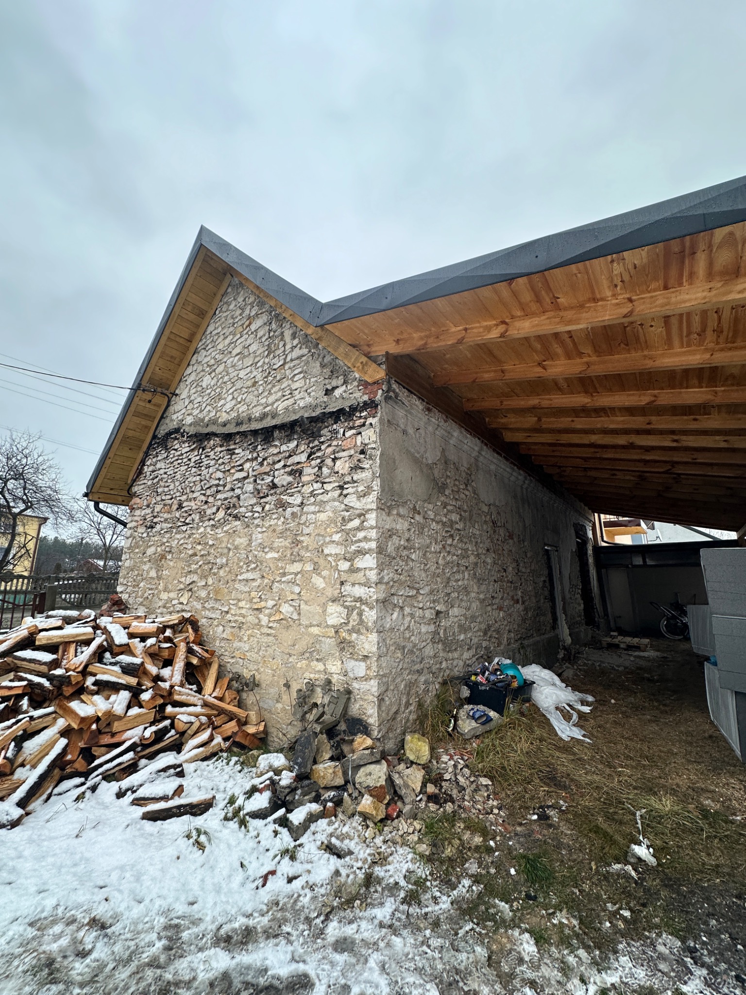 Kamienny budynek z drewnianą konstrukcją dachu w trakcie prac. Widoczne ślady demontażu starej elewacji i przygotowania do dalszych prac budowlanych. Stos drewna obok.