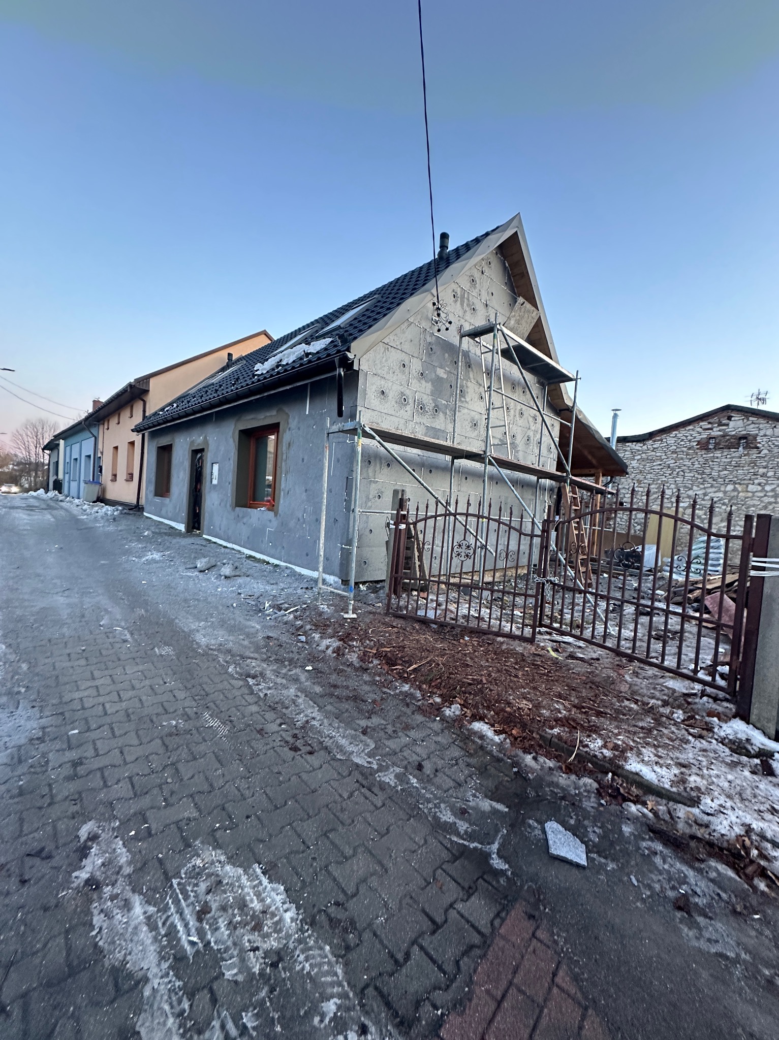 Dom w trakcie ocieplania, widoczne rusztowania i płyty izolacyjne na elewacji. Częściowo ośnieżony chodnik i brama w tle. Prace w toku, wczesny etap ocieplania.