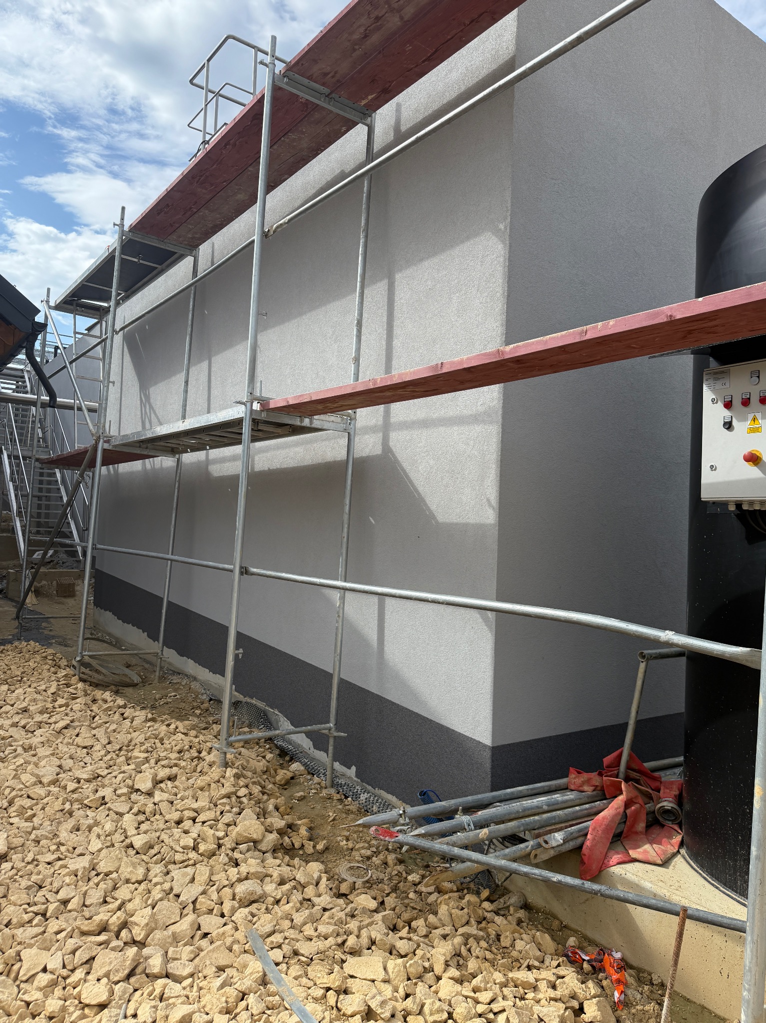 Ściana budynku w trakcie prac elewacyjnych, widoczne rusztowania i fragment ocieplonej, pomalowanej ściany z wykończonym cokołem. Na ziemi gruz i narzędzia.