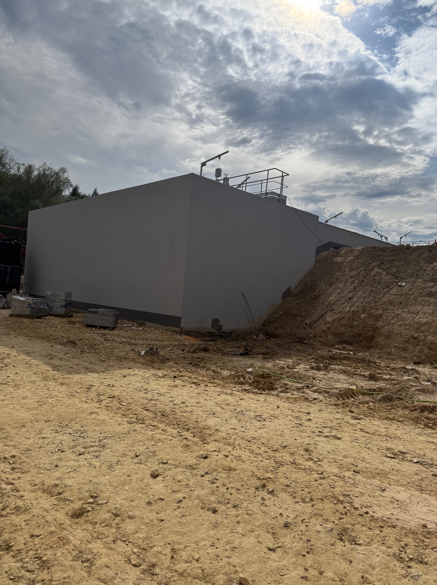 Szara, otynkowana ściana budynku w trakcie budowy, z widocznym nasypem ziemi i materiałami budowlanymi. Pochmurne niebo w tle. Widoczne elementy konstrukcyjne na dachu.