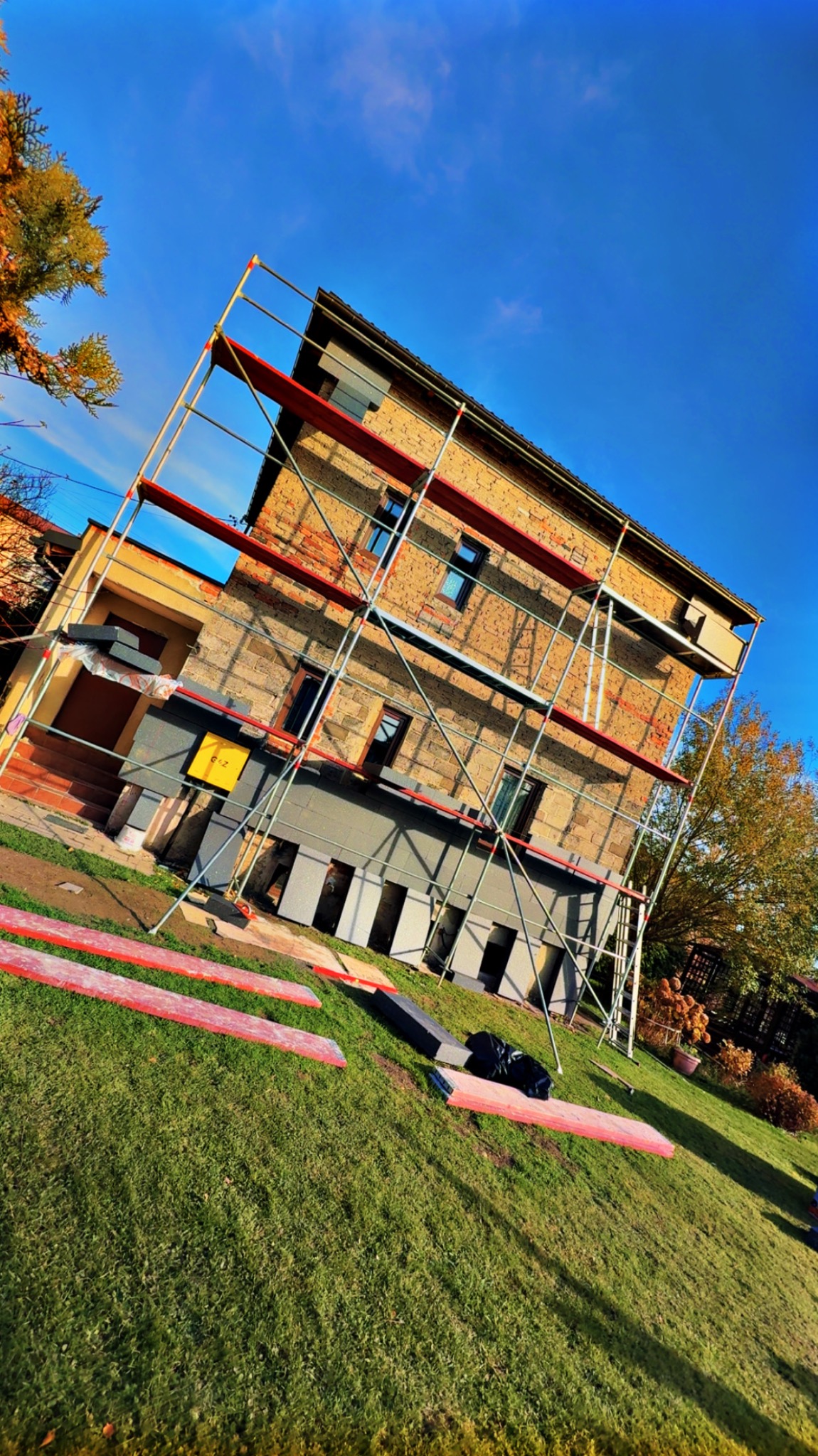 Dom z cegły w trakcie ocieplania, otoczony rusztowaniem. Częściowo ocieplona dolna partia elewacji, widoczne płyty izolacyjne na trawniku. Błękitne niebo w tle.