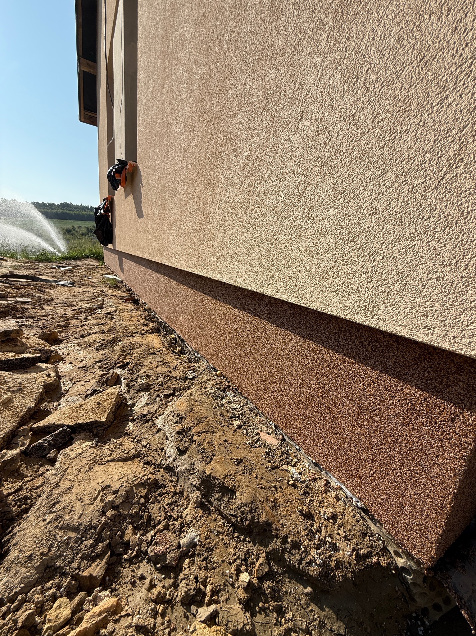 Wykończona elewacja budynku z widocznym cokołem w kolorze brązowym. W tle zraszacze nawadniające trawnik. Ujęcie z dołu, podkreślające strukturę elewacji.