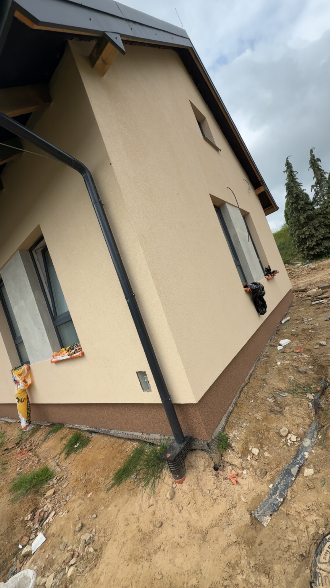 Otynkowana elewacja domu jednorodzinnego z brązowym cokołem, widoczne okna i rynna. Teren wokół domu w trakcie prac wykończeniowych.