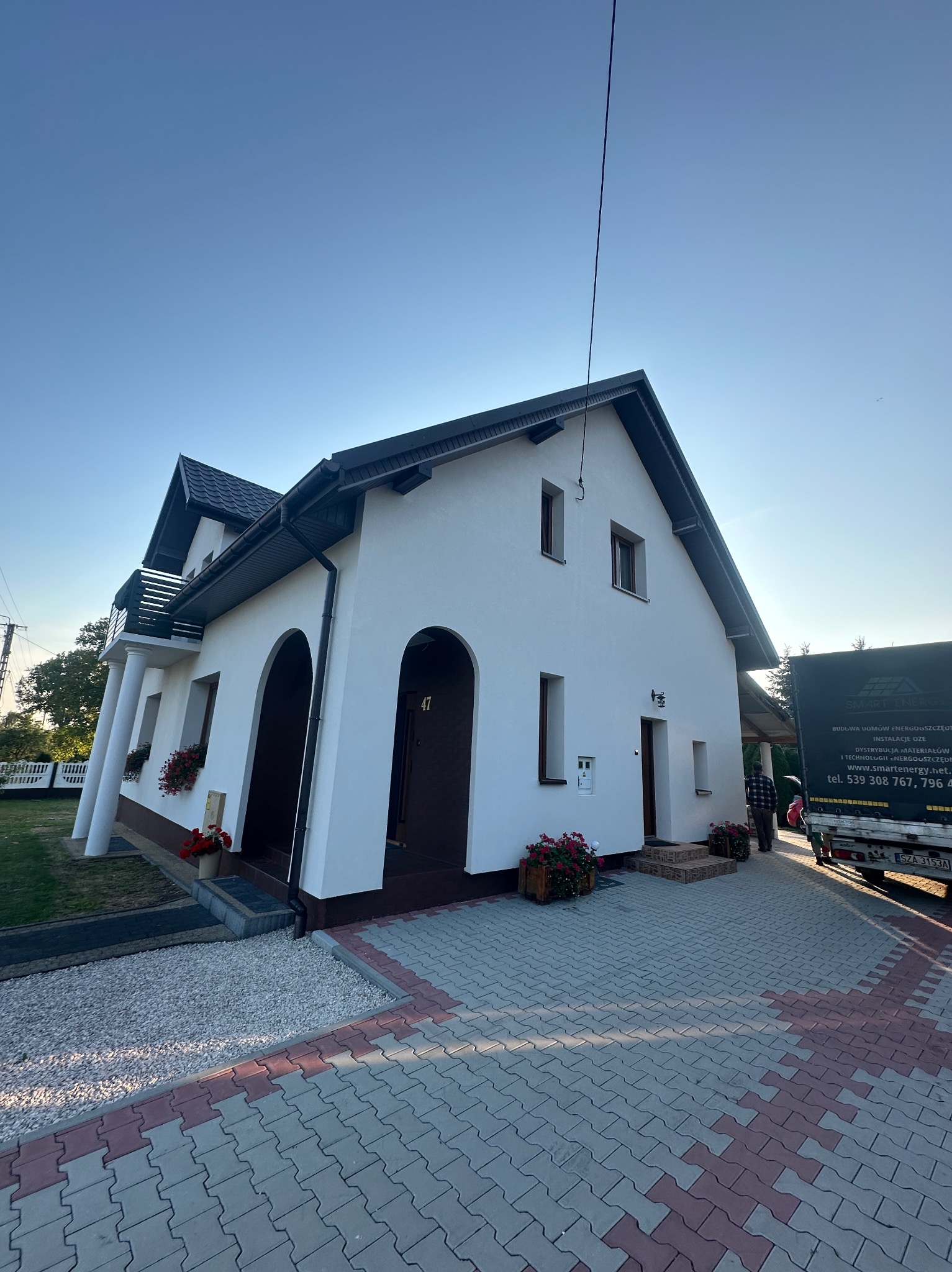 Biały dom jednorodzinny z ciemnym dachem i stolarką okienną, widoczny z poziomu chodnika z kostki. Na podjeździe stoi częściowo widoczny samochód dostawczy.