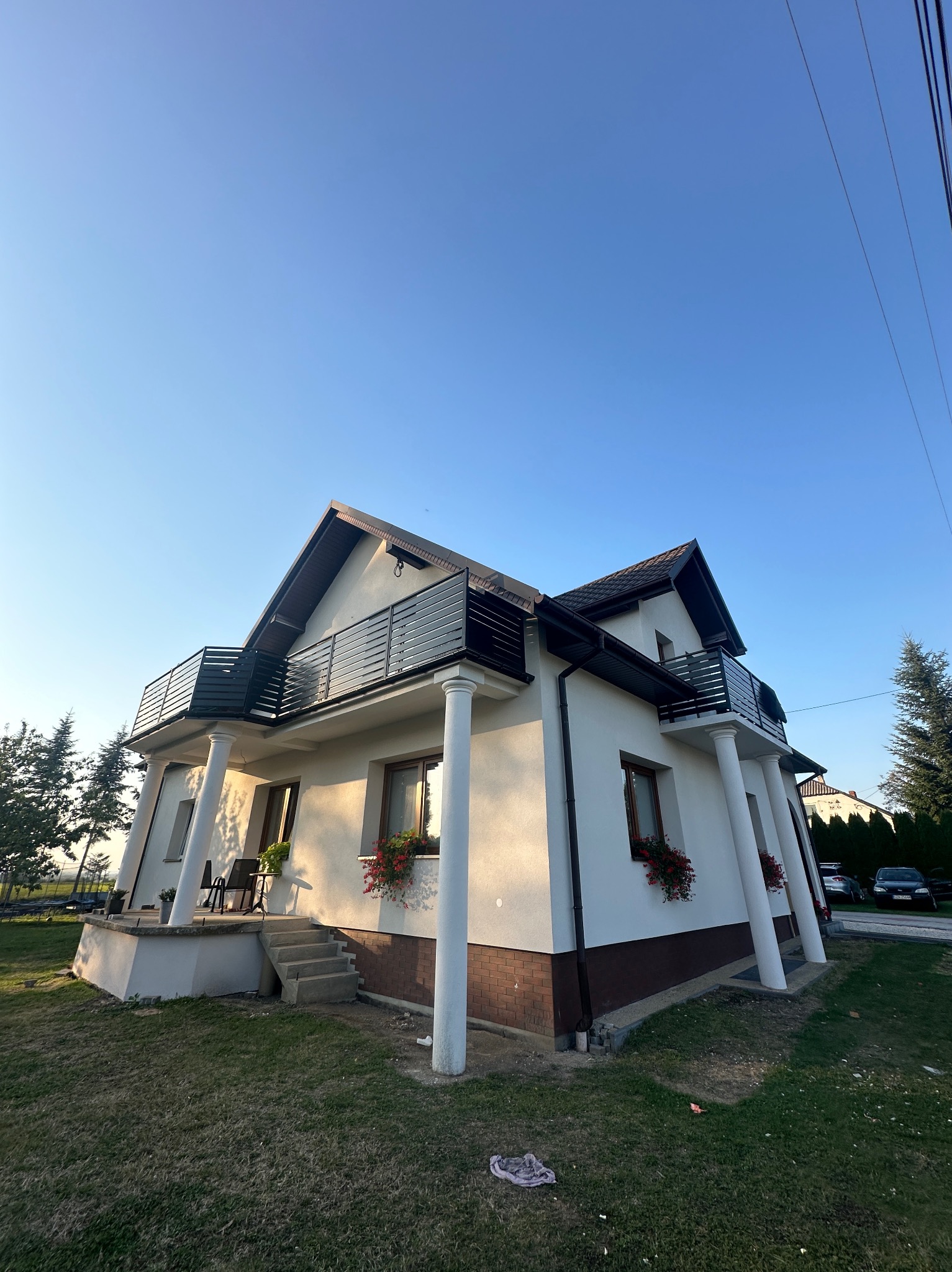 Dwupiętrowy dom z balkonami, kolumnami i kwiatami w oknach, sfotografowany z dołu pod kątem, na tle błękitnego nieba. Widoczne elementy elewacji i otoczenie domu.