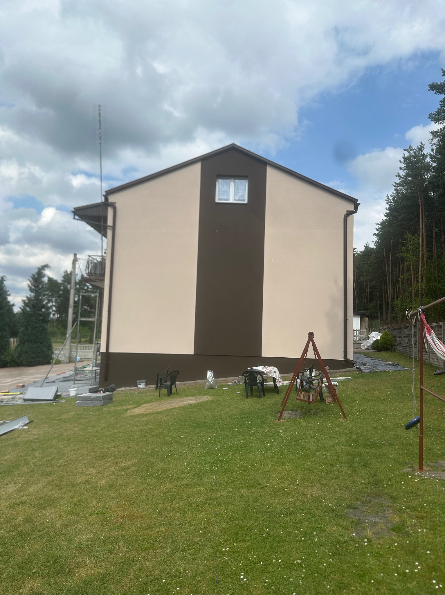 Dwukolorowa elewacja domu z beżowymi ścianami i brązowym pasem, widoczna z boku, z rusztowaniem i elementami placu budowy w tle, na tle zielonego trawnika.