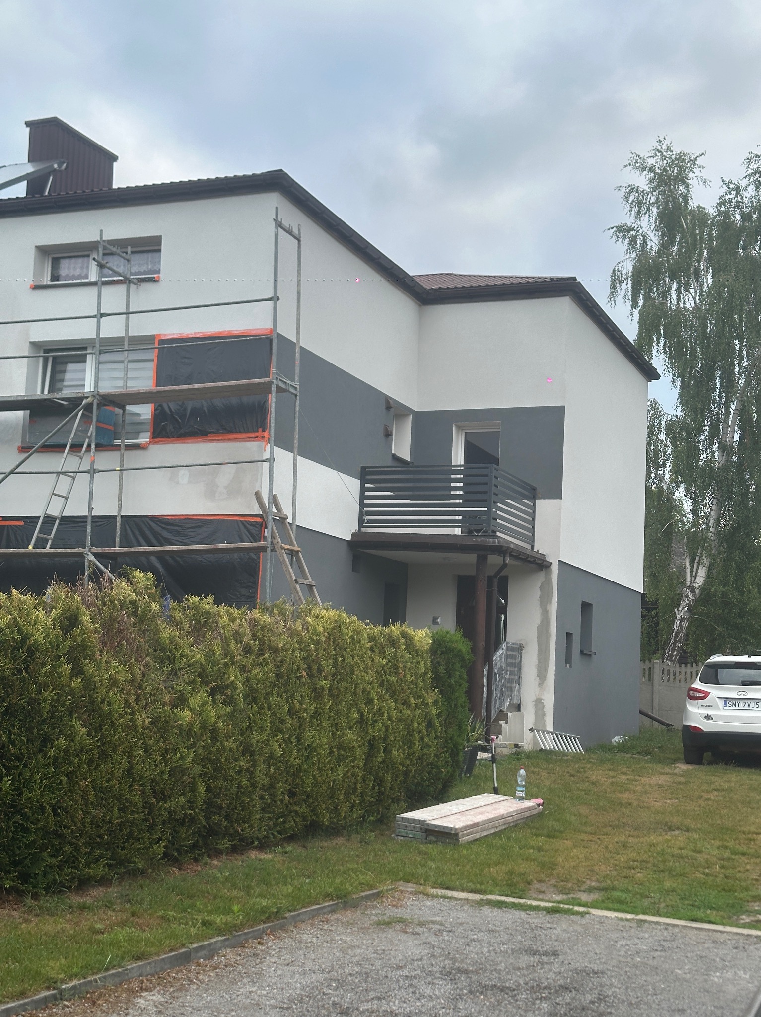 Dwukondygnacyjny dom z balkonem w trakcie malowania elewacji na szaro-biało. Widoczne rusztowania, zabezpieczenia okien folią i fragment ogrodu z samochodem.