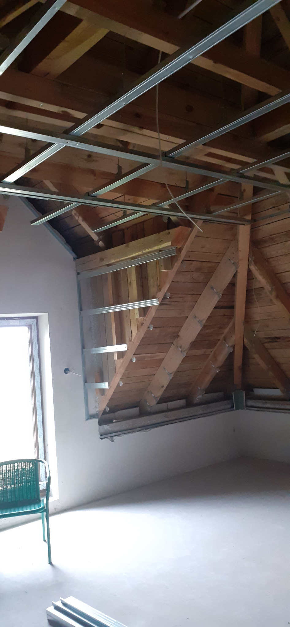 Montaż metalowej konstrukcji sufitu podwieszanego w poddaszu z drewnianą więźbą. Widoczny fragment okna i krzesło. Zdjęcie w trakcie prac.