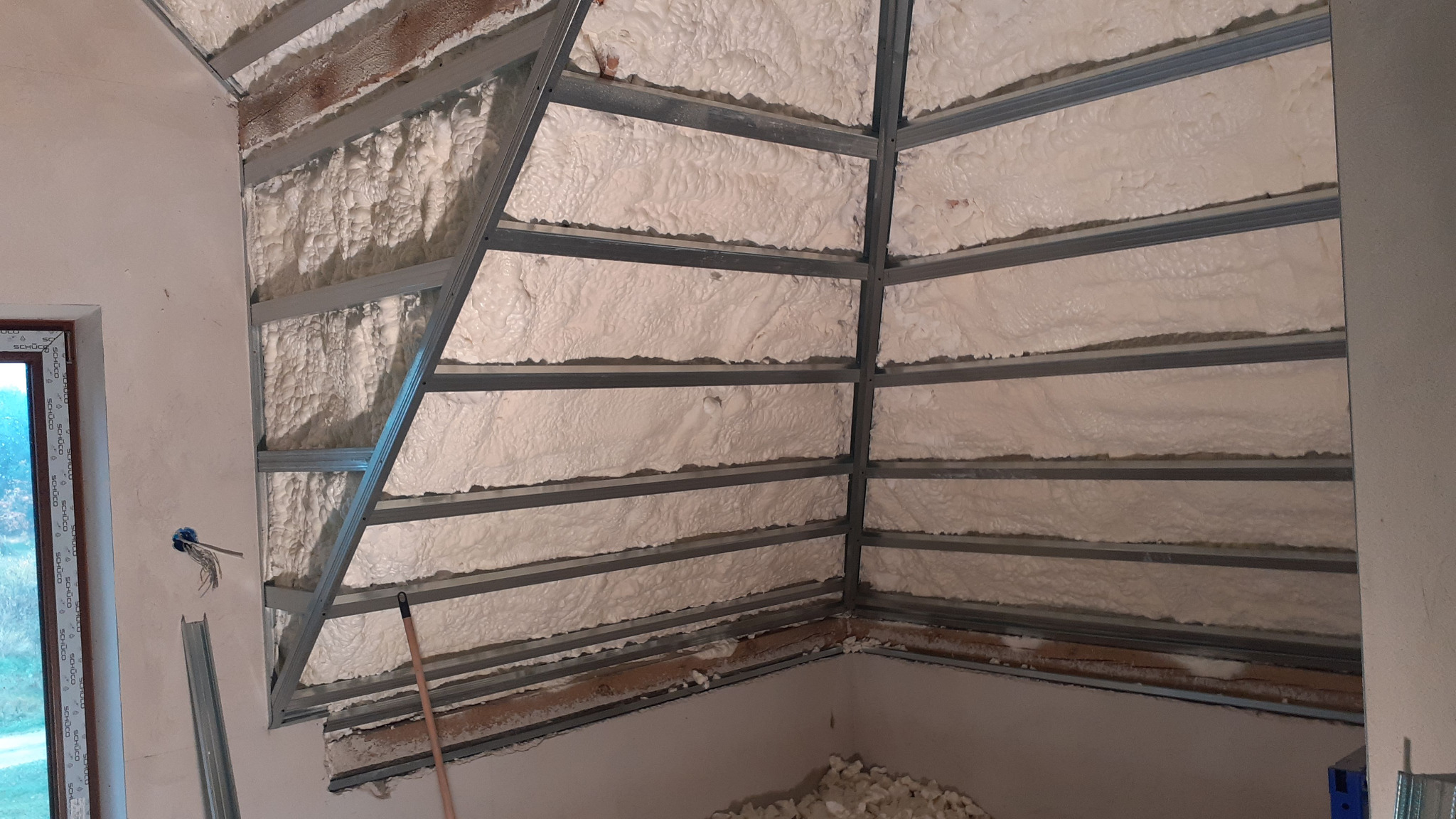 Skośna ściana poddasza w trakcie izolacji pianą PUR, widoczne metalowe profile konstrukcyjne. Fragment okna po lewej stronie. Praca w toku.
