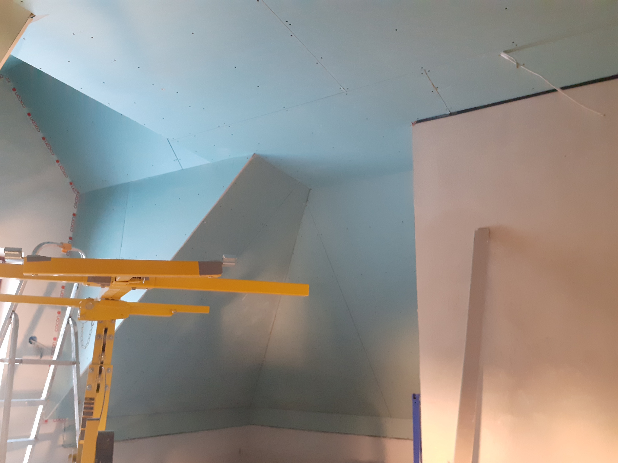 Montaż płyt gipsowo-kartonowych na poddaszu, widoczny podnośnik do płyt i fragment ściany. Praca w toku, sufit i ściany pokryte płytami, widoczne łączenia.