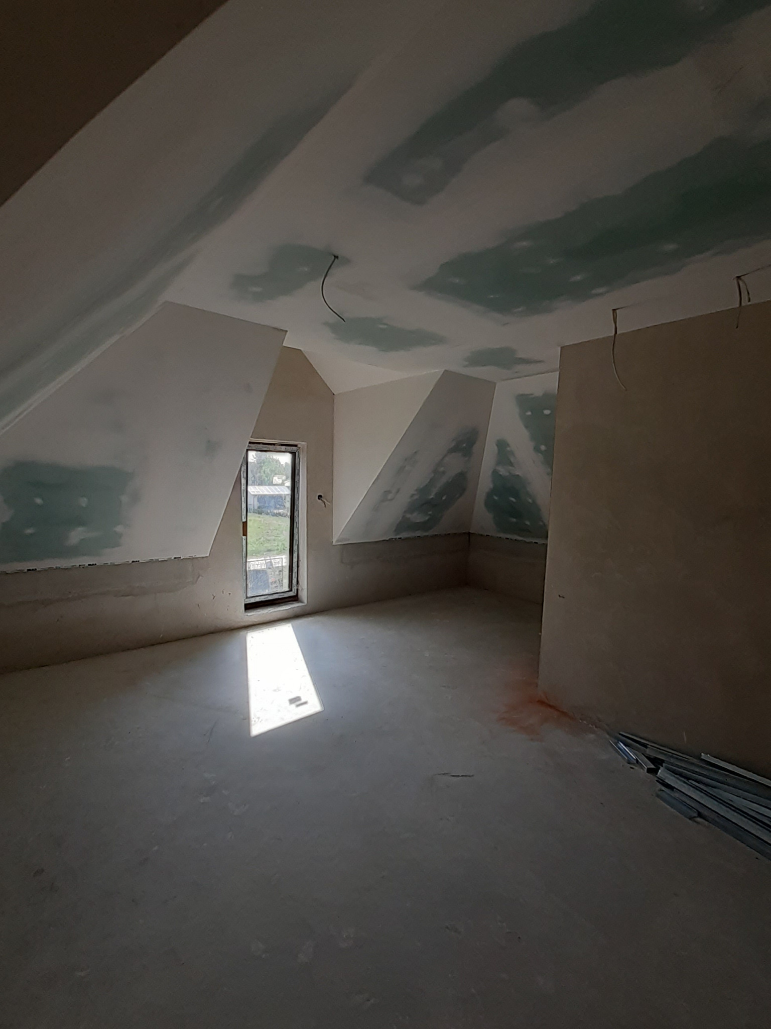 Pokój na poddaszu po szpachlowaniu i montażu płyt gipsowo-kartonowych, widoczne łączenia płyt. Wpadające światło przez okno. Metalowe profile na podłodze.