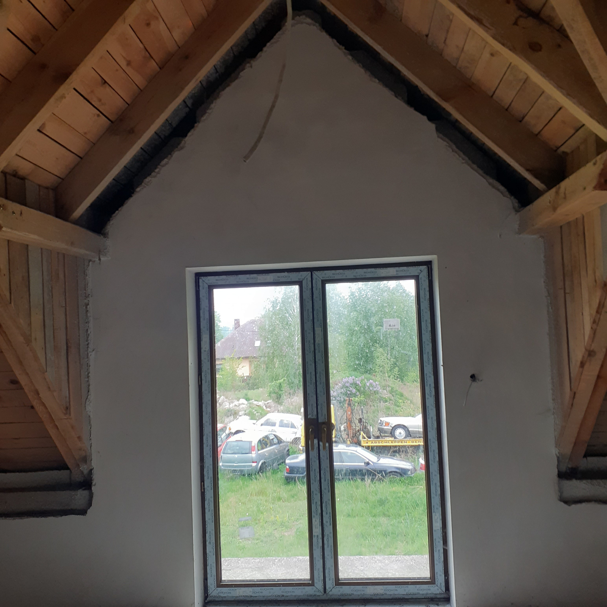 Surowa ściana poddasza ze szpachlowaniem, widoczne drewniane elementy konstrukcyjne dachu i okno z widokiem na zewnątrz. Etap prac wykończeniowych.