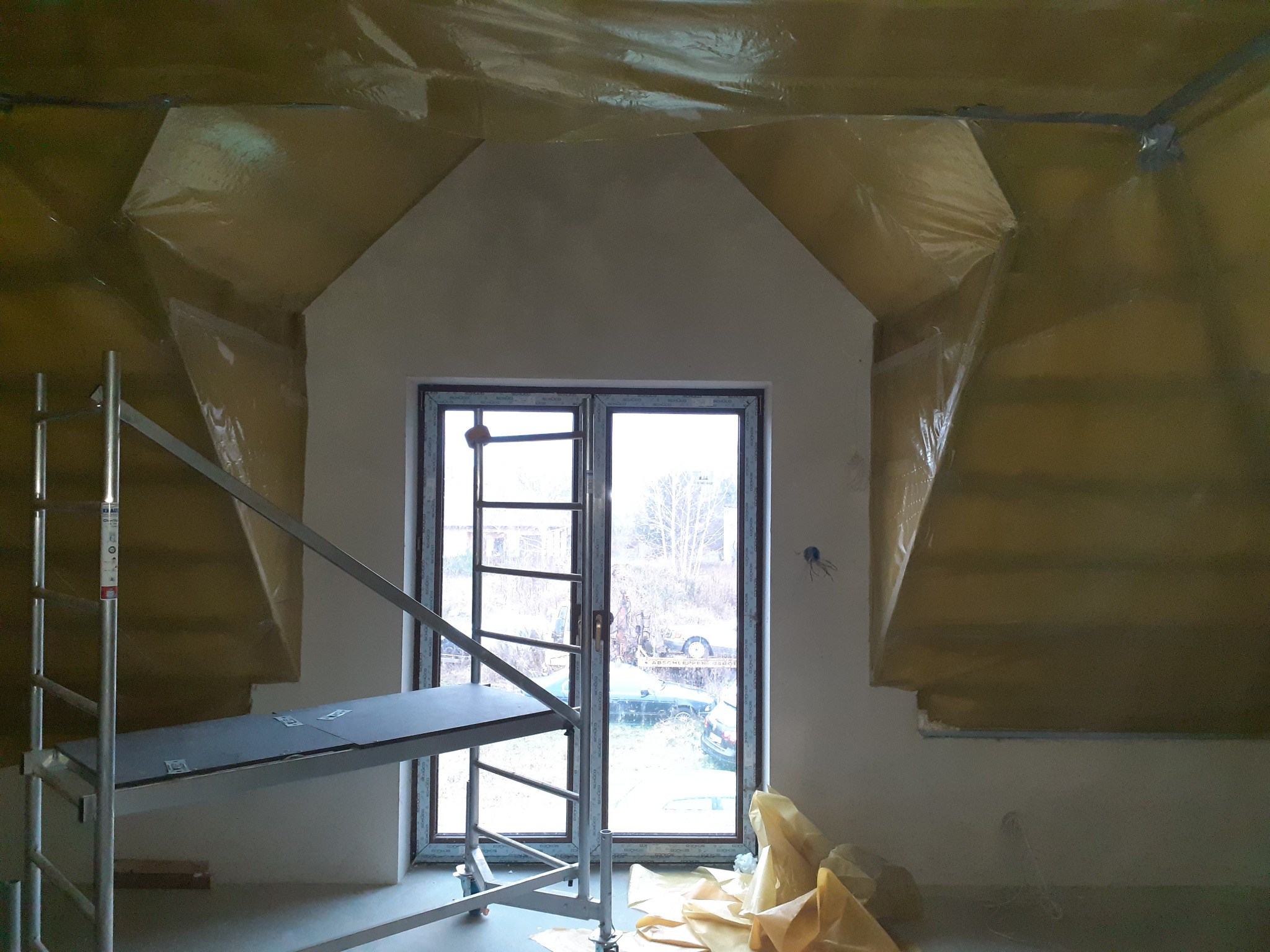 Pokój na poddaszu w trakcie remontu: ściany po szpachlowaniu, zabezpieczone folią, widoczne rusztowanie i okno z widokiem na zewnątrz. Prace wykończeniowe w toku.