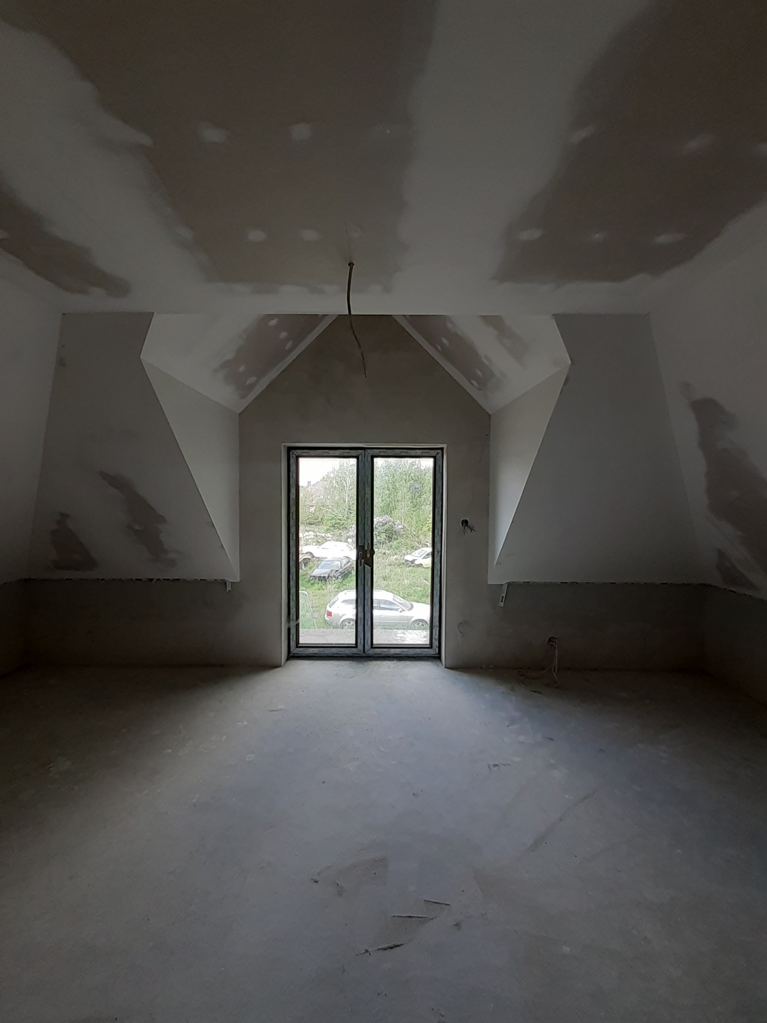 Surowe wnętrze poddasza z oknem balkonowym, ściany i sufit po szpachlowaniu, przygotowane do malowania. Widoczne niedoskonałości i surowy beton na podłodze.