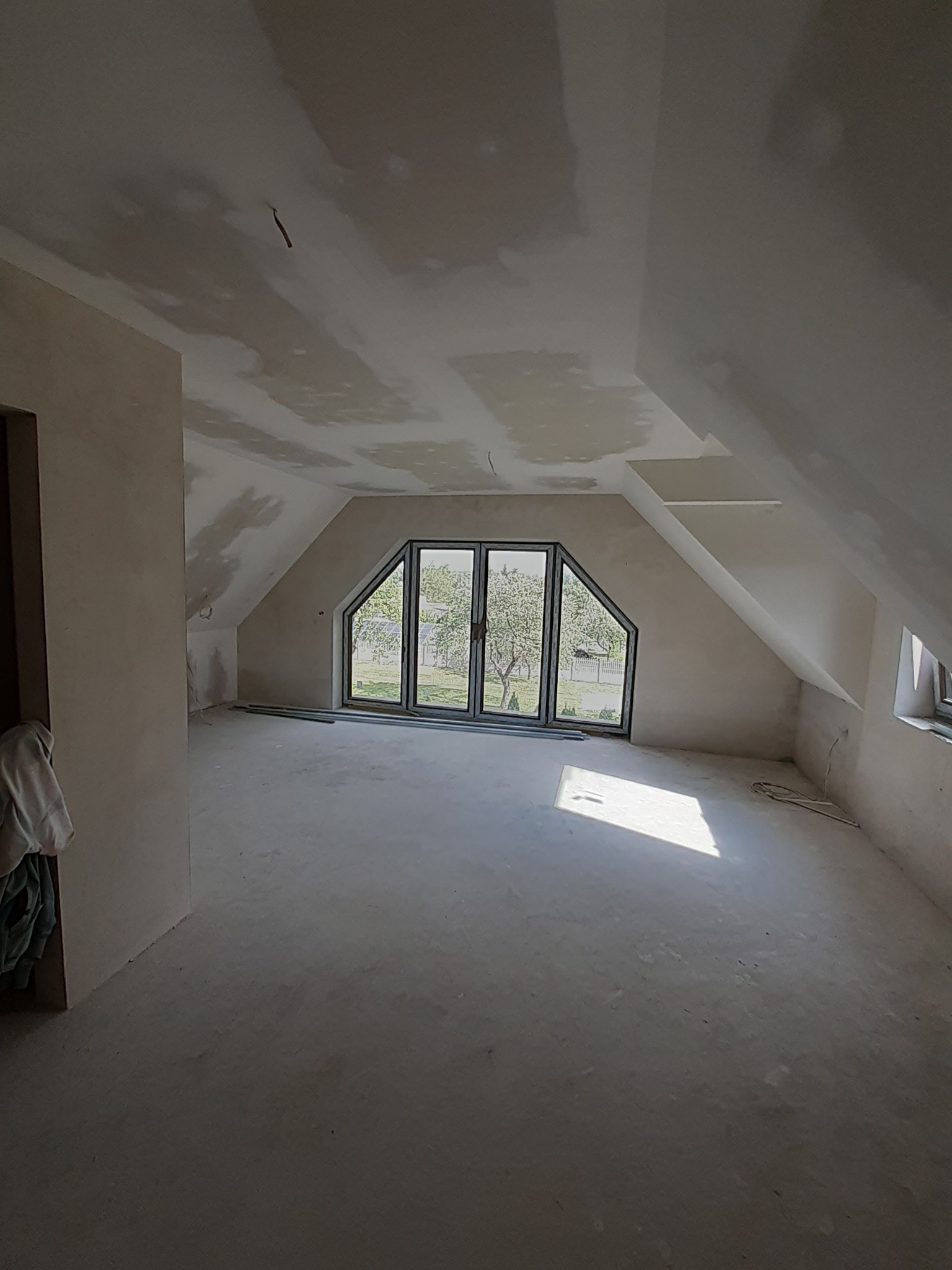Poddasze w trakcie wykończenia: szpachlowane ściany, sufit z płyt karton-gips, okno z widokiem na ogród. Widoczne ślady prac wykończeniowych.