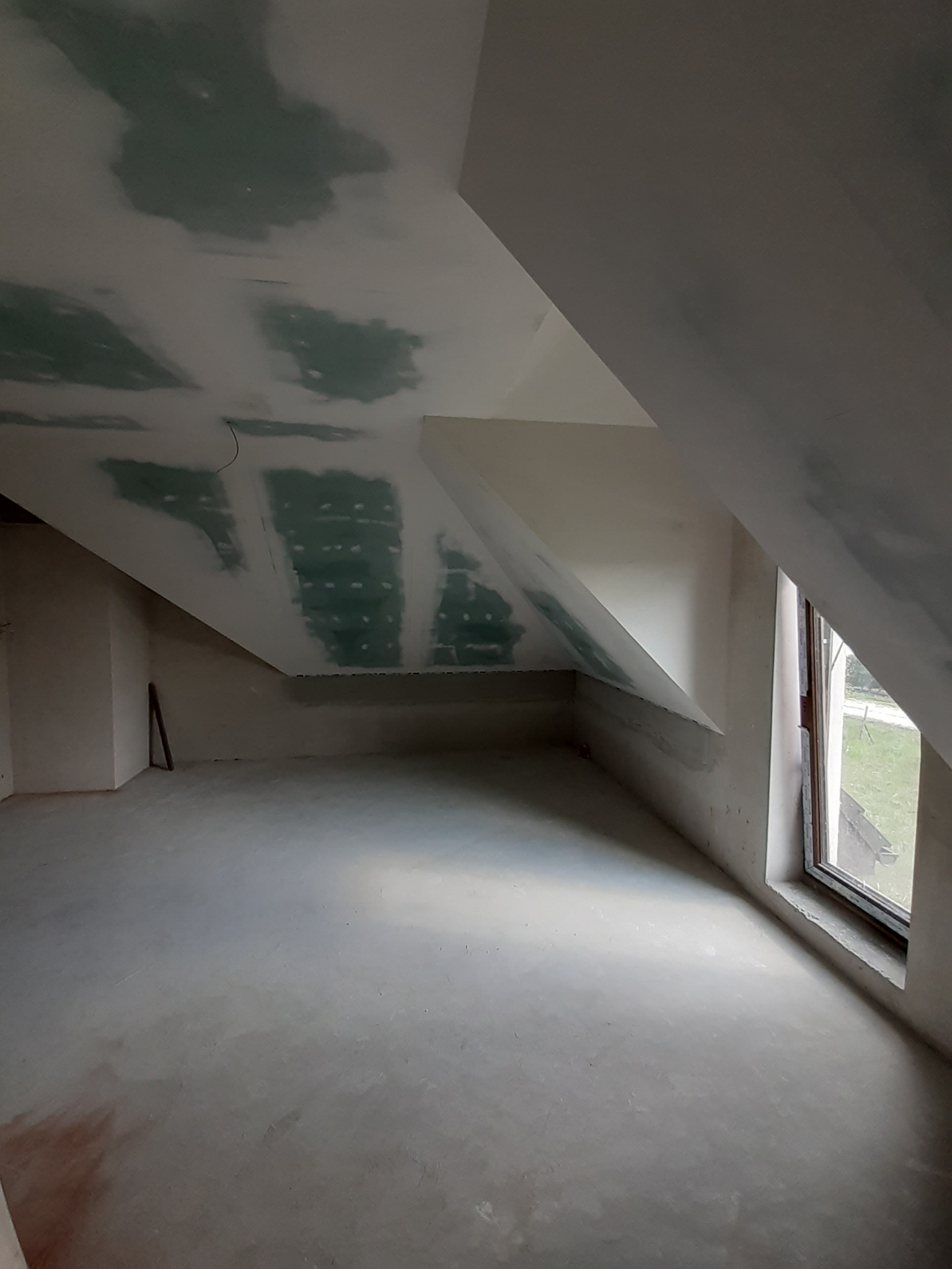 Poddasze w trakcie szpachlowania: sufit z płyt karton-gips, częściowo pokryty szpachlą, okno w ścianie szczytowej, surowa podłoga. Widok na wnętrze.