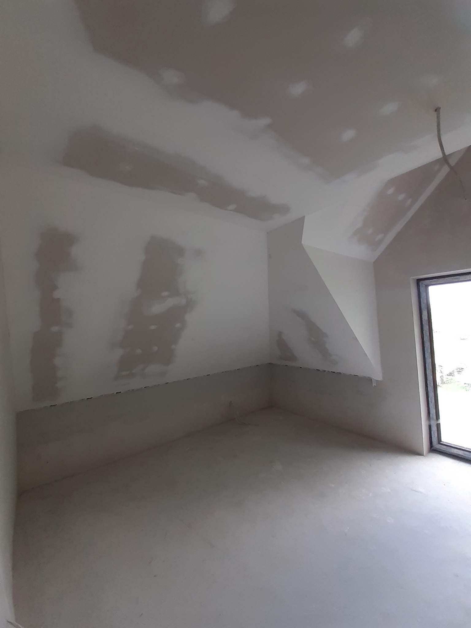 Poddasze po szpachlowaniu, widoczne ślady po łatach na ścianach i suficie. Ściany w kolorze jasnego beżu, okno w czarnej ramie. W tle widoczne niebo.