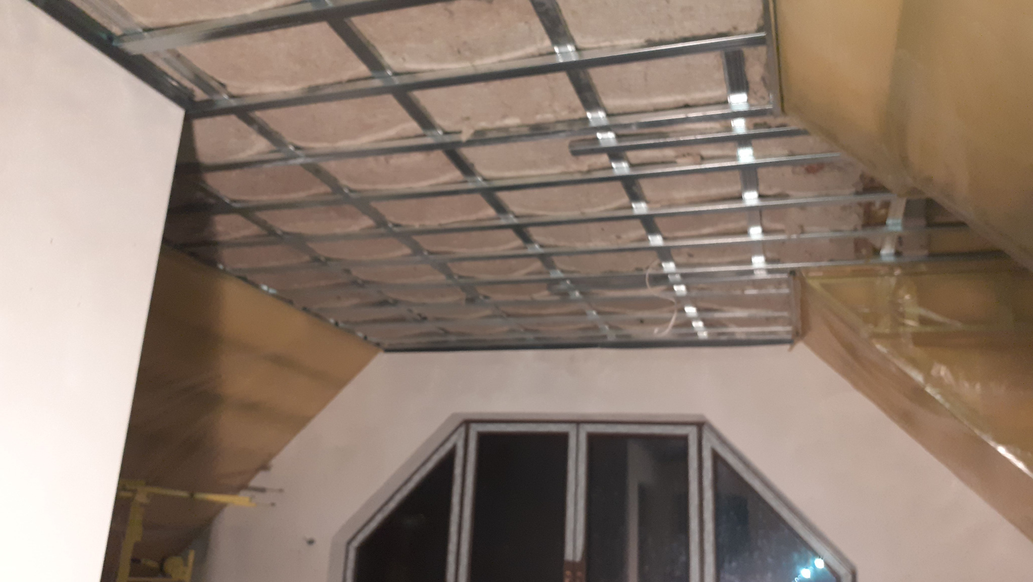 Montaż izolacji sufitu podwieszanego z wełny mineralnej w pomieszczeniu ze skośnym dachem i oknem. Widoczne profile metalowe i folia ochronna na ścianach.