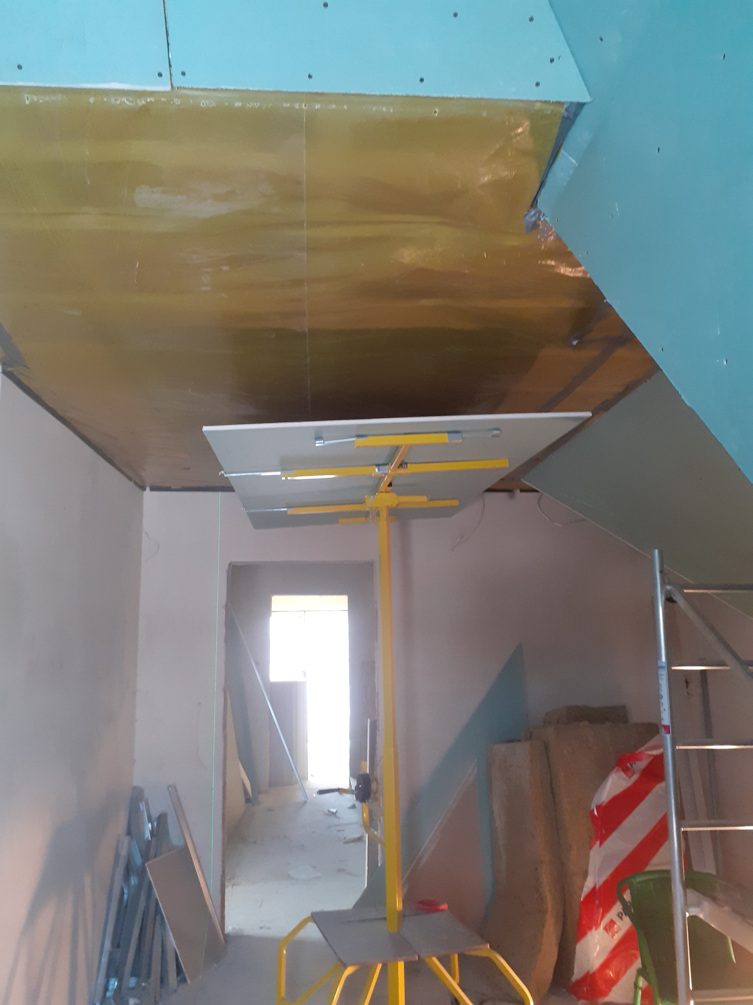 Montaż izolacji i płyt karton-gips na suficie poddasza. Widoczny podnośnik do płyt, folia izolacyjna i częściowo zamontowane płyty. Prace wykończeniowe w trakcie.