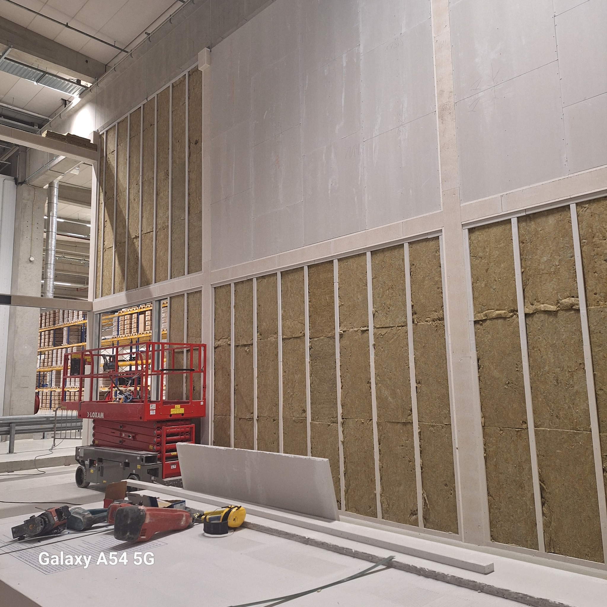 Montaż izolacji z wełny mineralnej w metalowym stelażu, częściowo zakrytej płytami gipsowo-kartonowymi. Widoczny podnośnik i narzędzia na podłodze w hali.