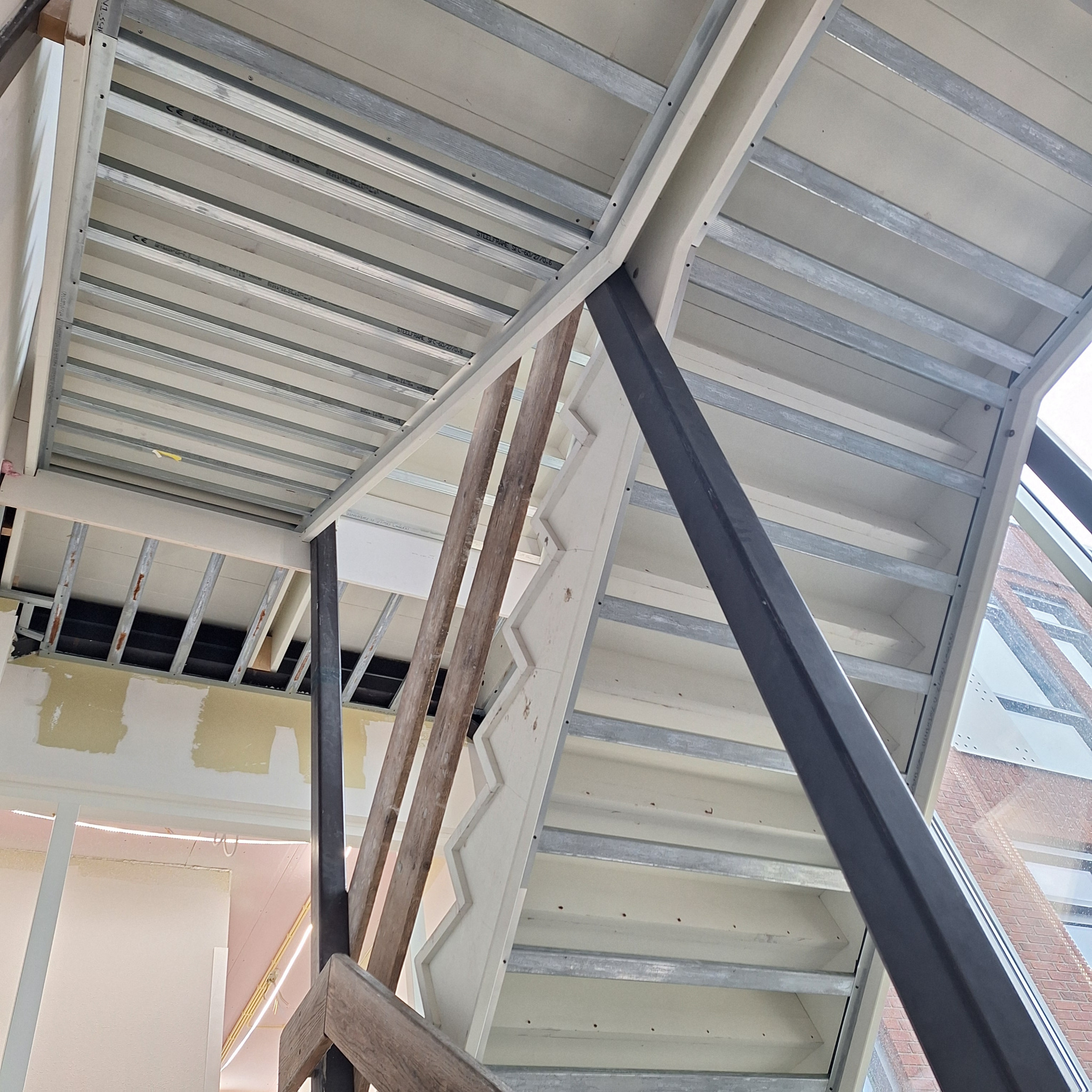 Sufit podwieszany z metalową konstrukcją i fragmentem schodów w trakcie montażu. Widoczne elementy konstrukcyjne i płyty gipsowe. W tle fragment budynku przez okno.