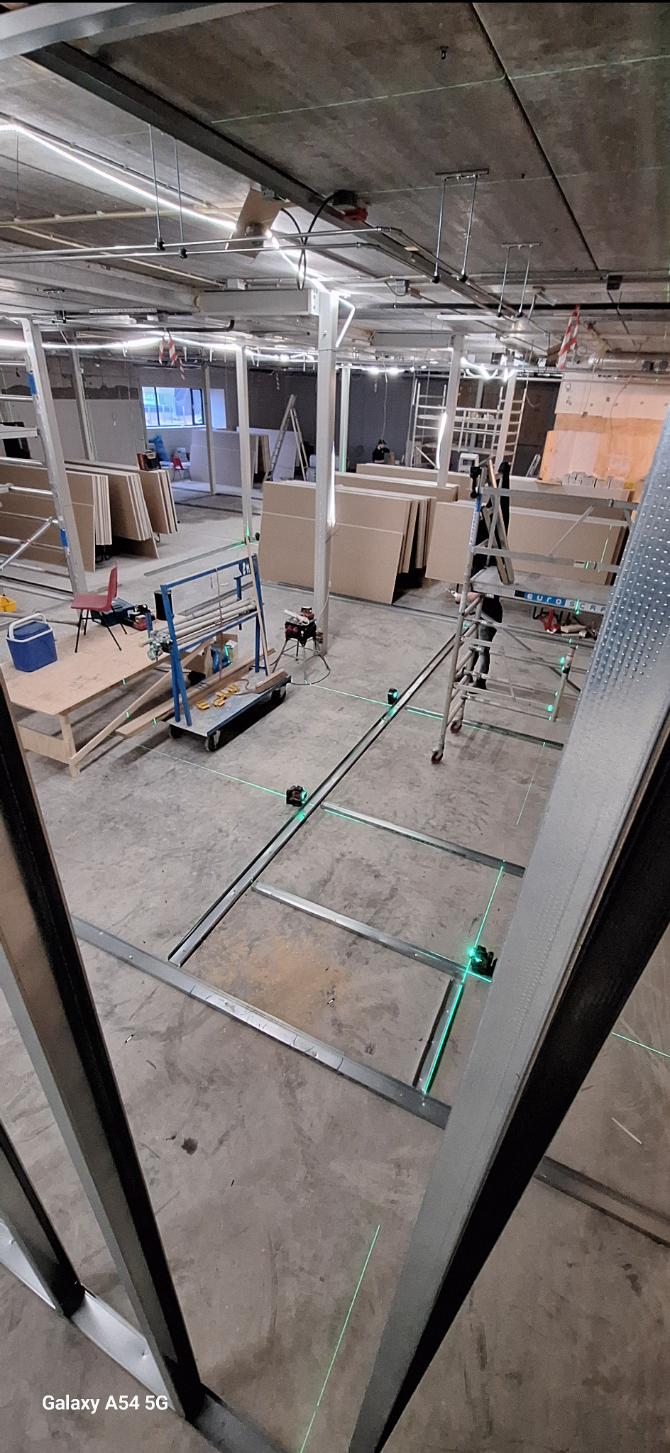 Montaż konstrukcji z profili stalowych, wyznaczanie poziomu laserem krzyżowym. W tle płyty karton-gips i drabina. Widok z góry na surowy stan wnętrza.