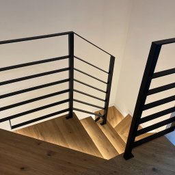 Michał Bogdański Boformi - Nowoczesne schody z drewnianymi stopniami i czarną, minimalistyczną balustradą. Ujęcie z góry, podkreślające geometrię i oświetlenie stopni.