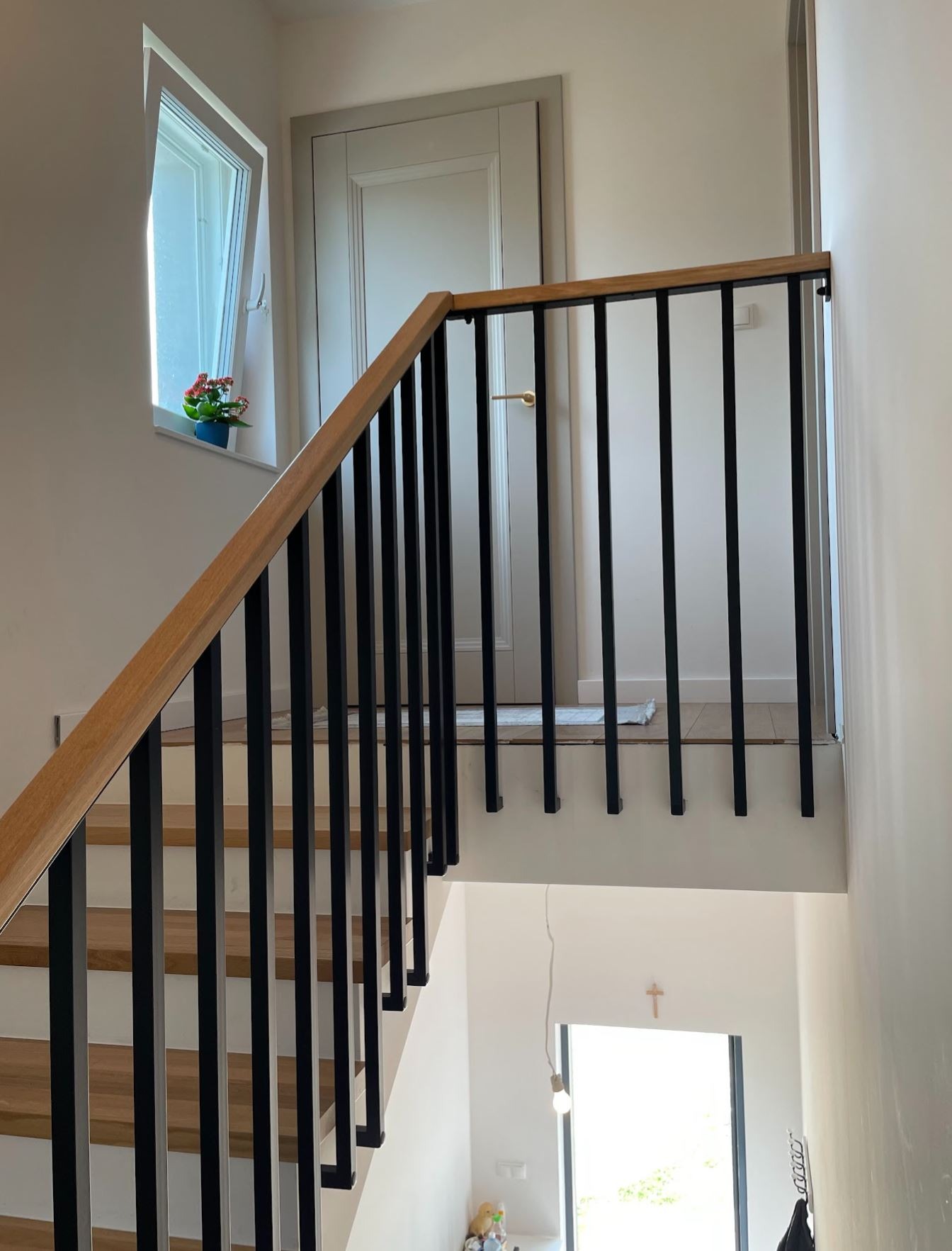 Nowoczesne schody z drewnianymi stopniami i czarną balustradą z pionowych prętów. Widok z dołu, w tle okno z kwiatami i drzwi.