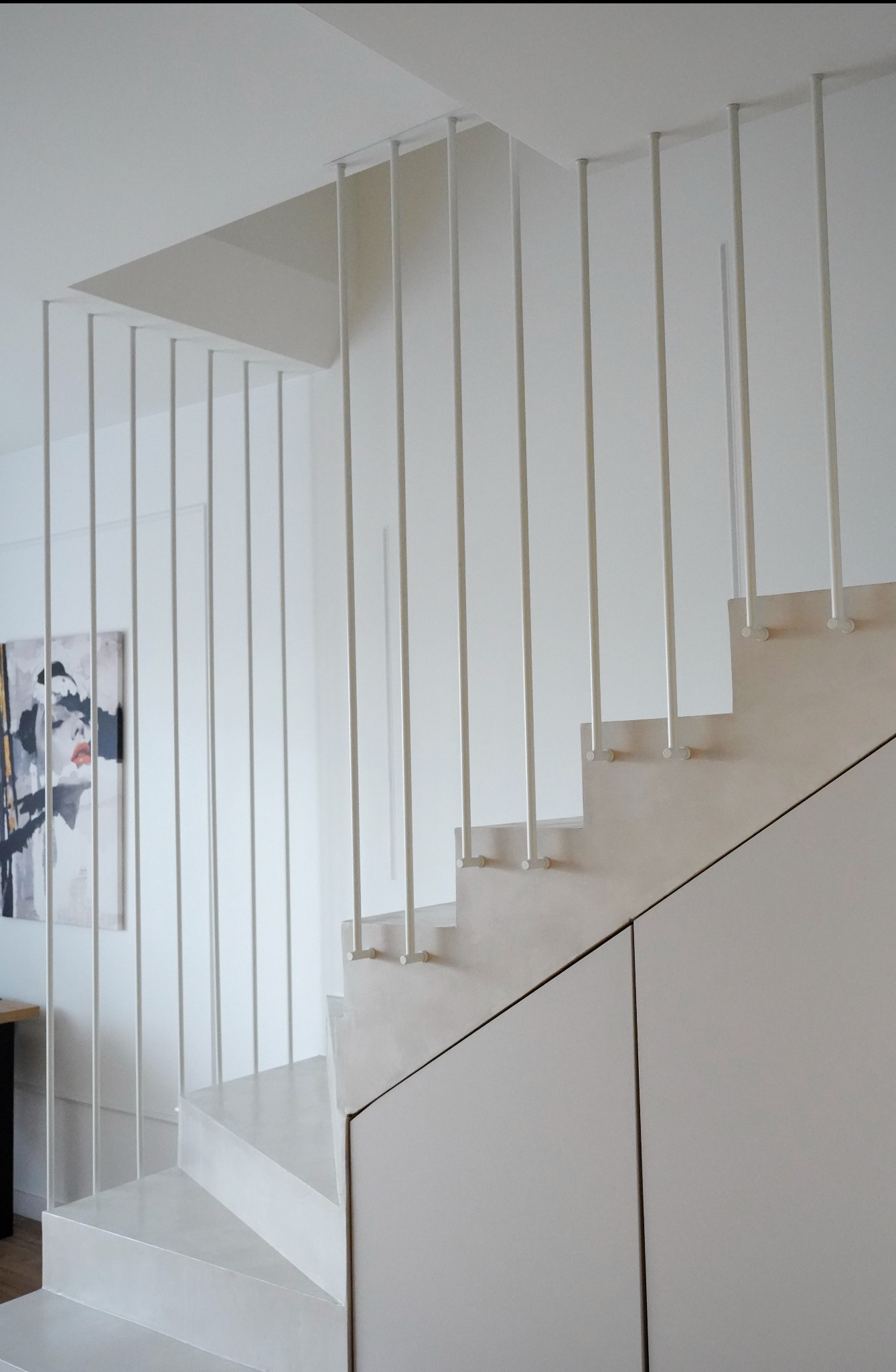 Minimalistyczne schody z białą balustradą z cienkich prętów, zintegrowane z zabudową szafkową pod schodami. Na ścianie widoczny abstrakcyjny obraz.